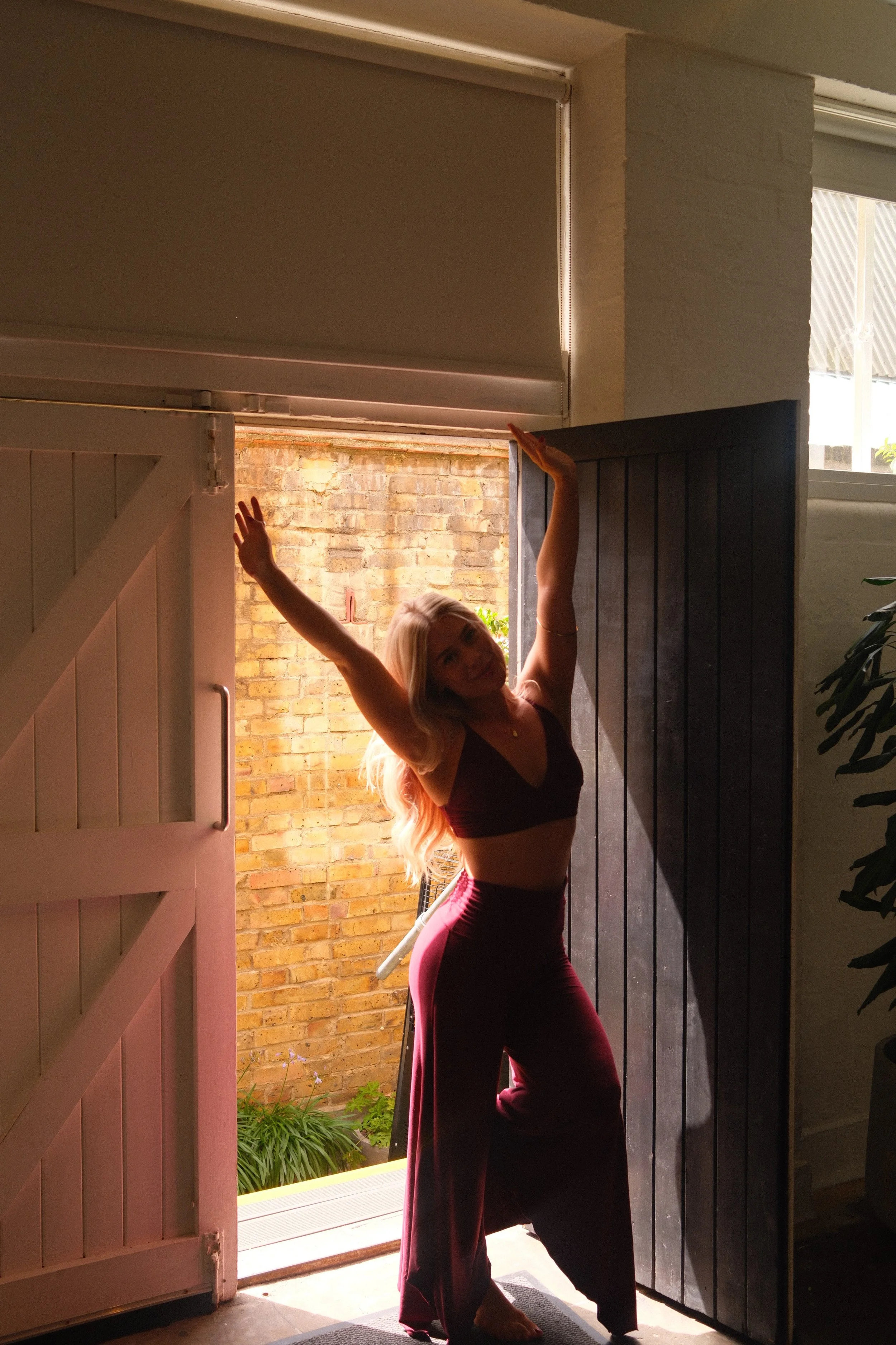 A woman with long blonde hair standing in a doorway, stretching her arms above her head. She is wearing a dark crop top and maroon wide-leg pants, smiling at the camera. The door is open to an outdoor space with a brick wall and greenery visible in the background.
