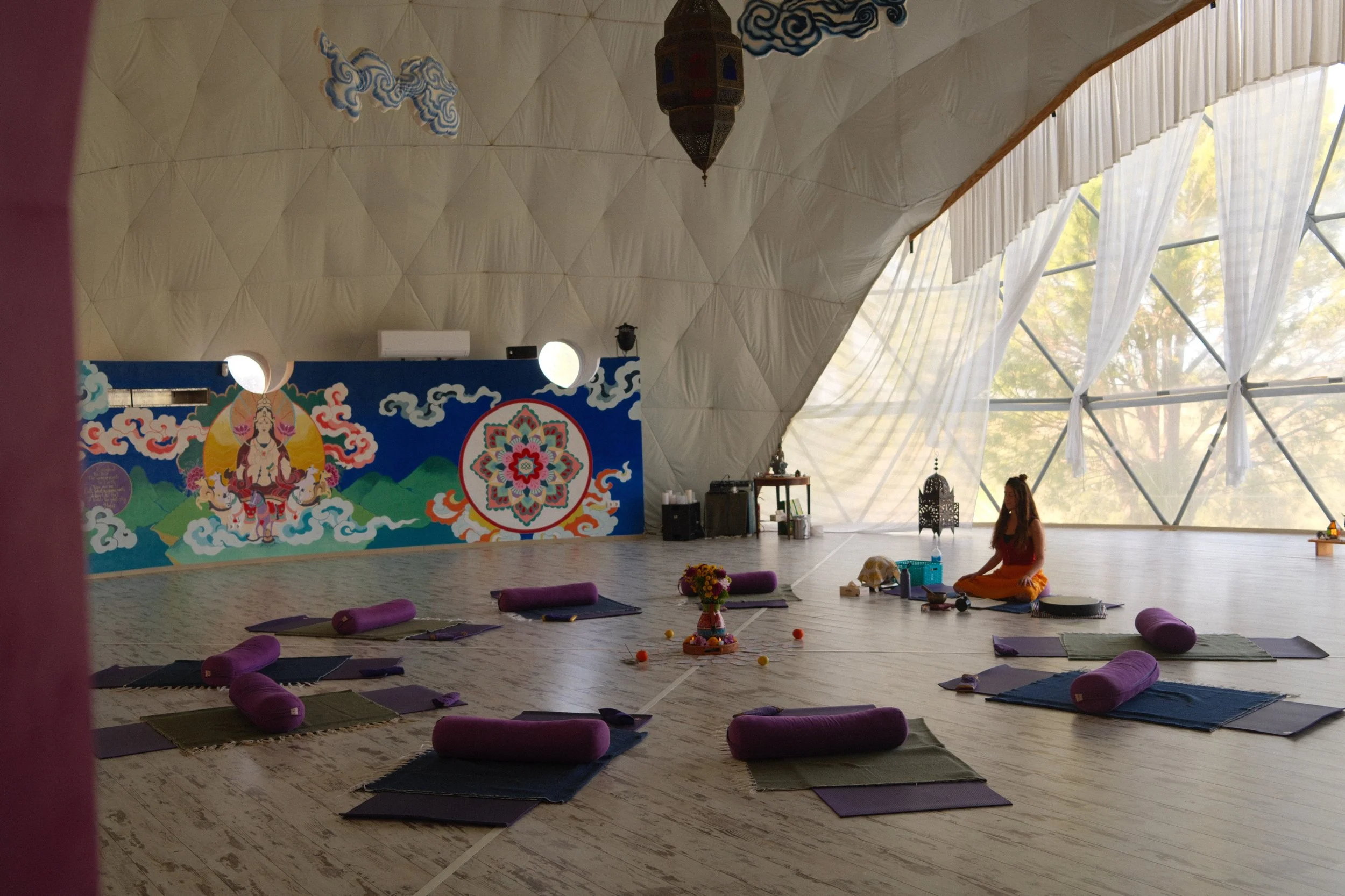 Interior of a yoga or meditation studio with a semi-dome ceiling and large windows with white curtains. A colorful mural of a goddess and abstract patterns is on the back wall. Yoga mats with purple bolsters are arranged in a circle on the floor, with a woman sitting cross-legged in the background, surrounded by various yoga props and decorative items.