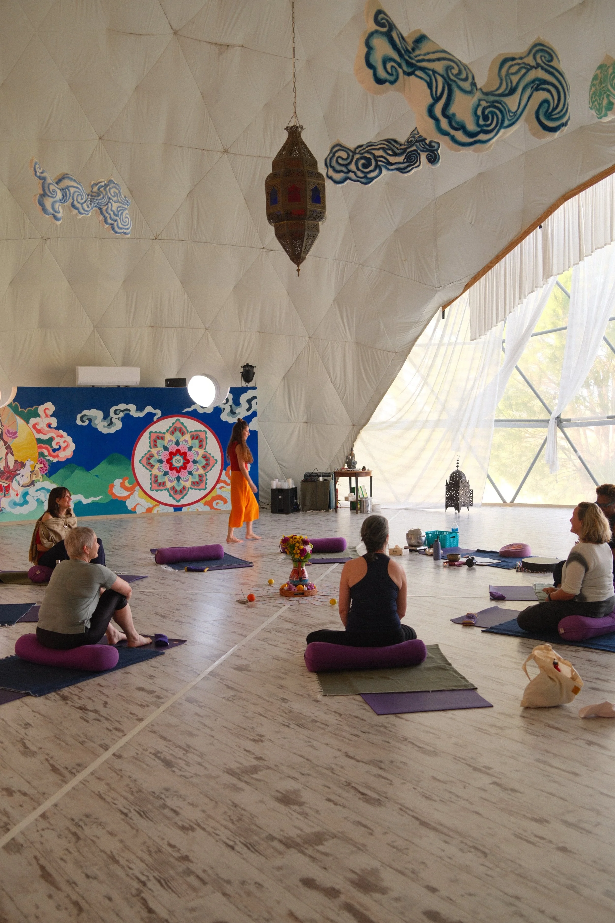 A group of people participating in a yoga or meditation class in a spacious, decorated studio with natural light, colorful wall art, and yoga mats arranged on the wooden floor.