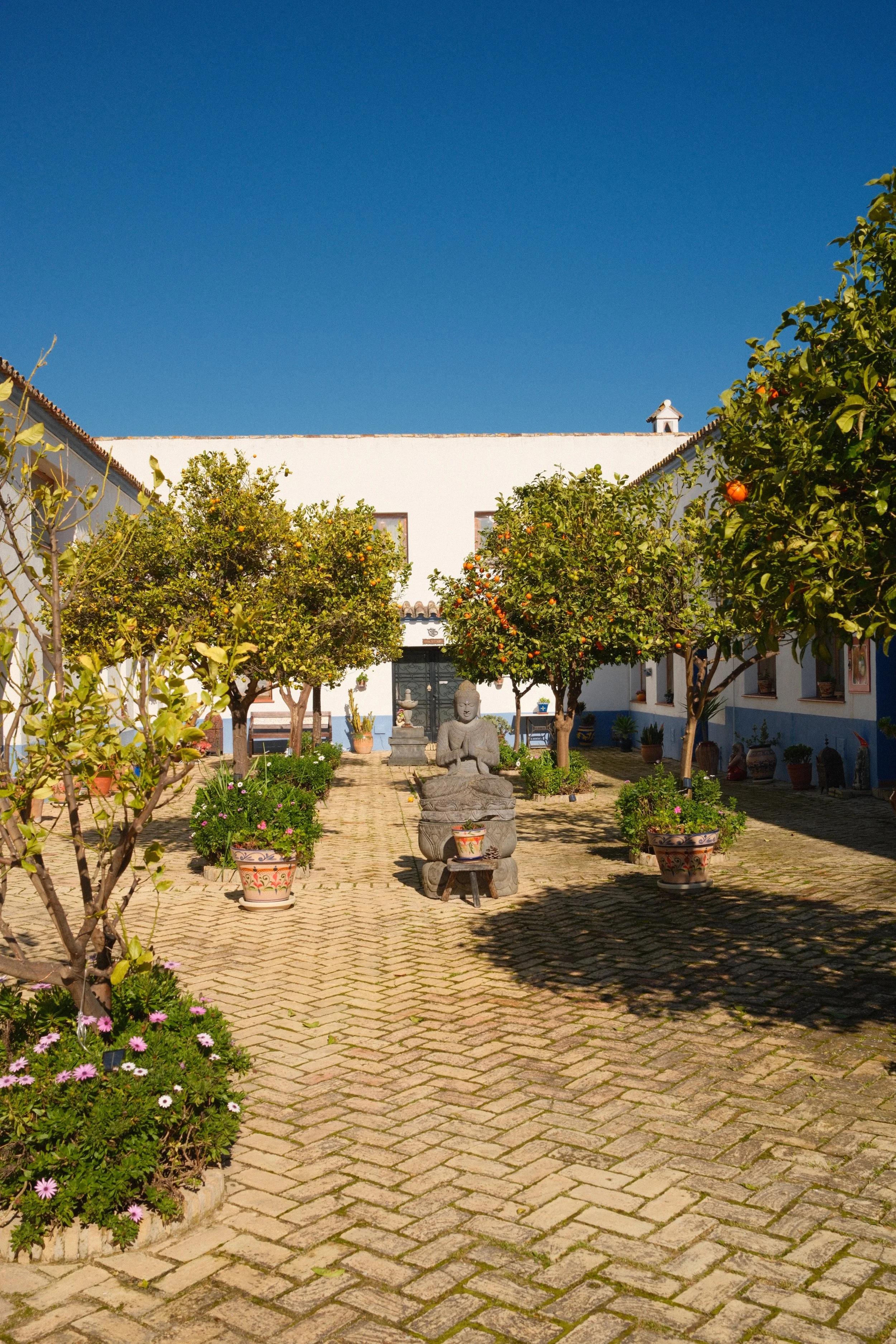 A courtyard with a brick paver floor, orange trees, and potted plants surrounding a stone Buddha statue. Homecoming yoga and creative retreat in Andalusia Spain with Meala Soul