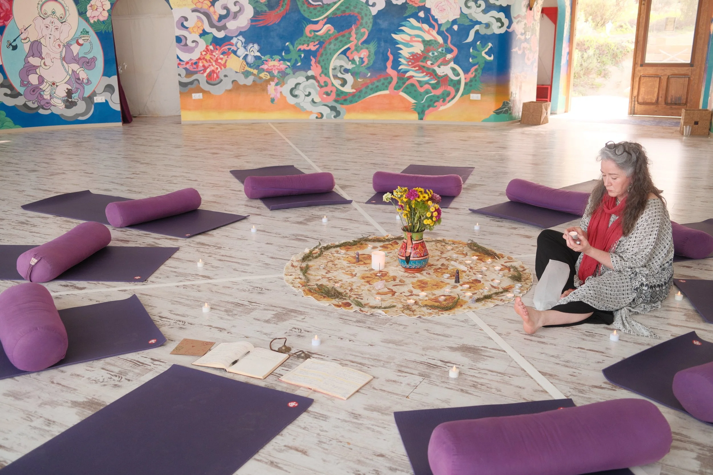 A woman sitting on the floor in a meditation room, surrounded by yoga mats and bolsters. She appears to be preparing for a meditation session, with a floral centerpiece, candles, and books on the floor.