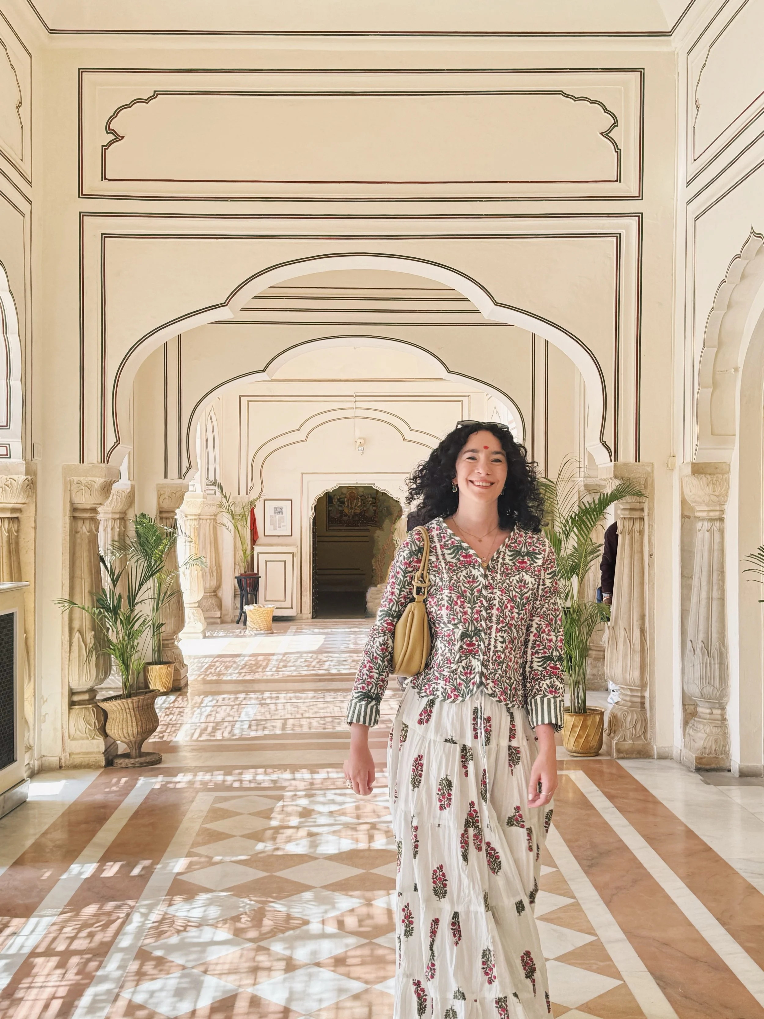 A smiling woman with curly black hair, dressed in a floral shirt and wide-legged, floral-printed pants, stands inside a brightly lit, ornate corridor with arches, potted plants, and intricate wall designs. Meala Soul India retreat jaipur yoga retreat
