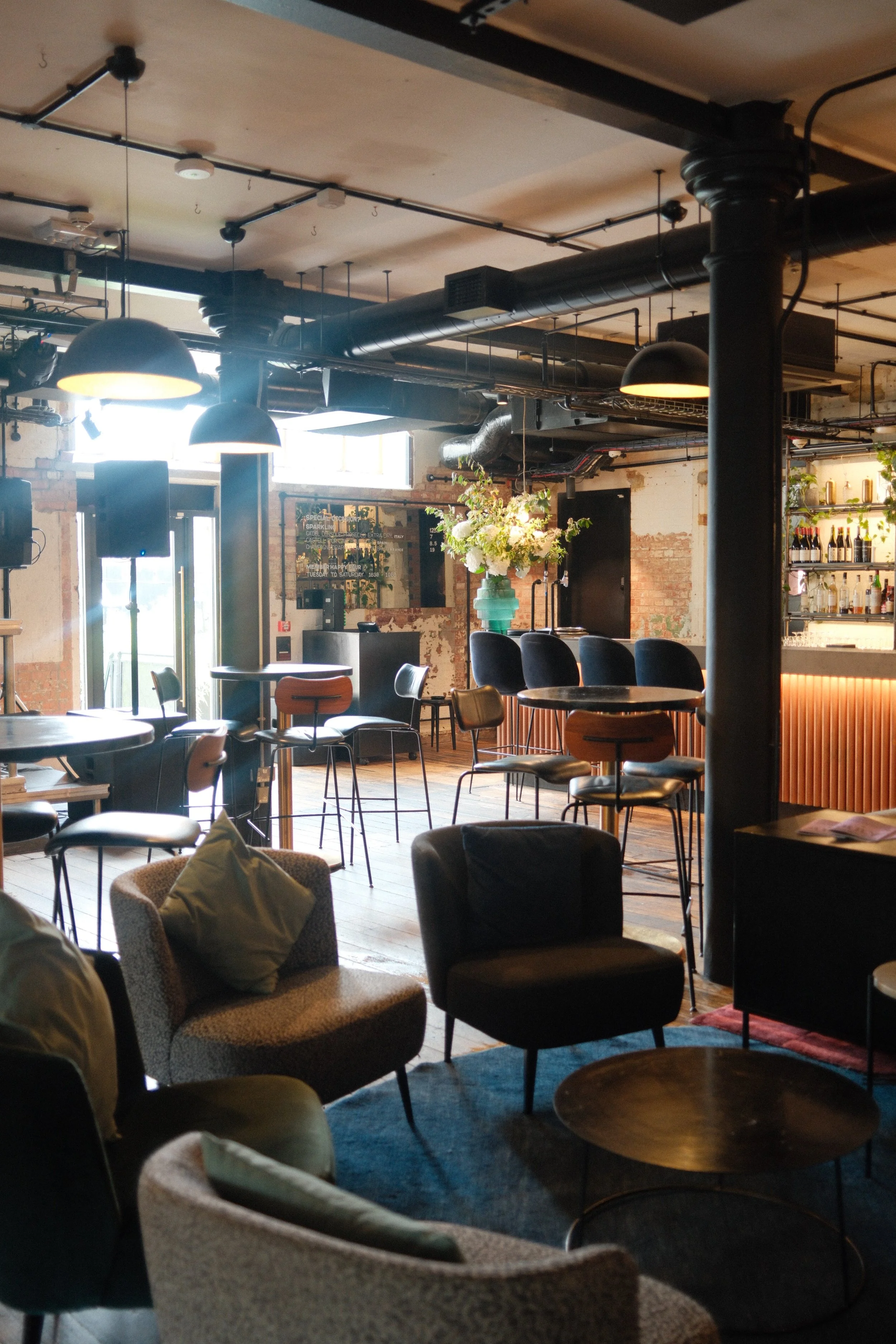 Interior of a modern cafe or bar with hanging black pendant lights, exposed ductwork, a bar area with bottles, a statue with flowers, and various chairs and tables.