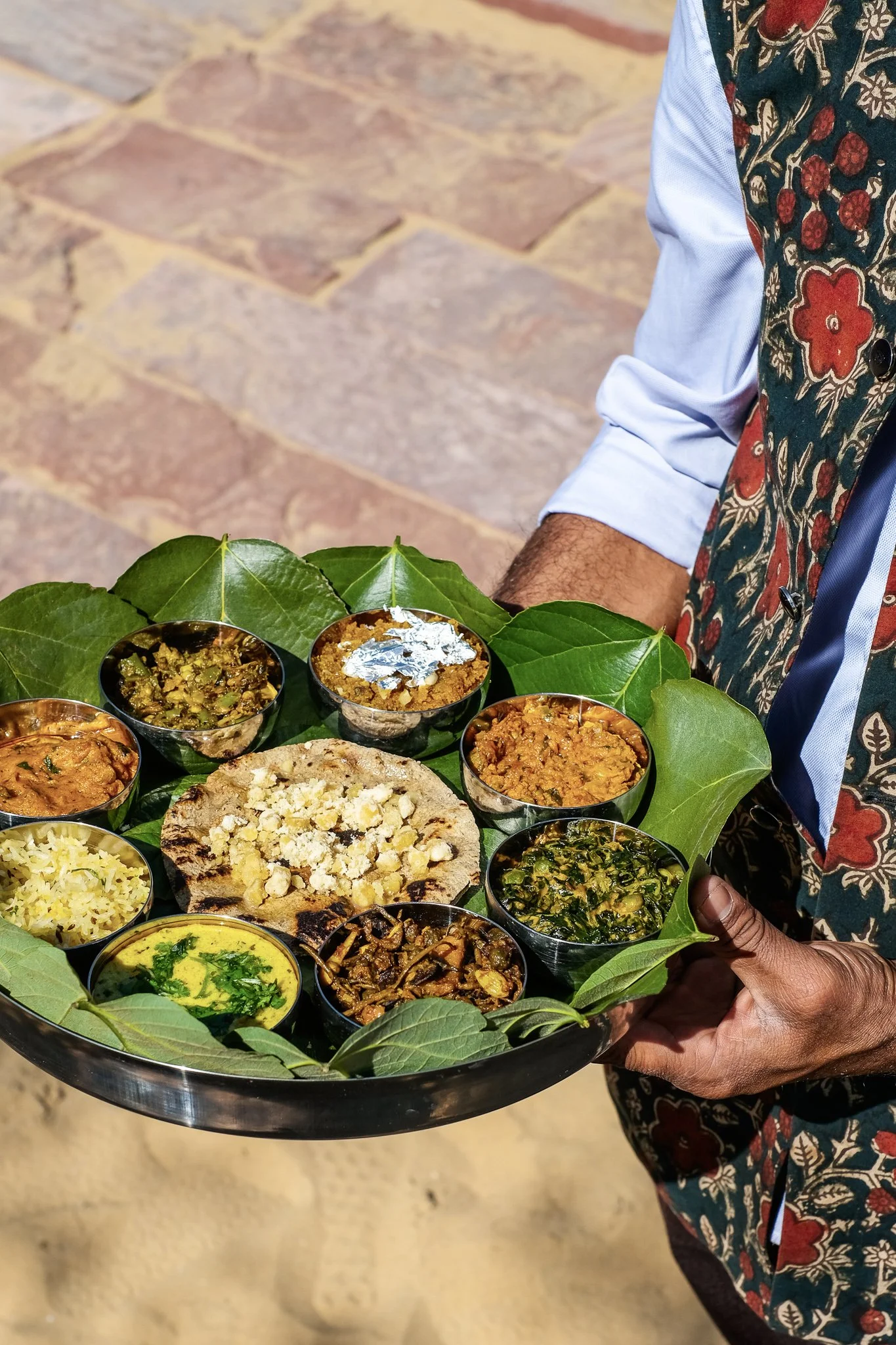 A person holding a large tray of various Indian foods, including small bowls of different curries and rice, with a naan bread in the center, decorated with green leaves.  Meala Soul India retreat jaipur yoga retreat