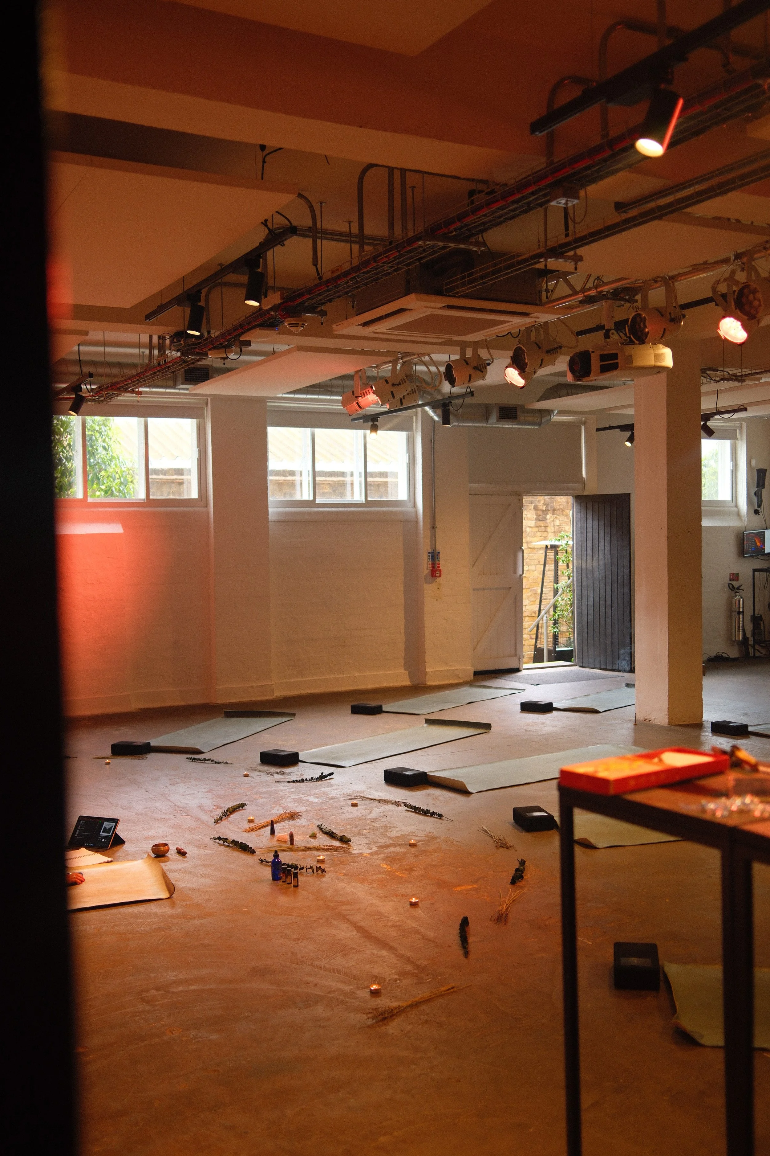 Empty indoor yoga or meditation studio with floor mats, candles, and decorative objects arranged on the floor, with large windows and natural light.