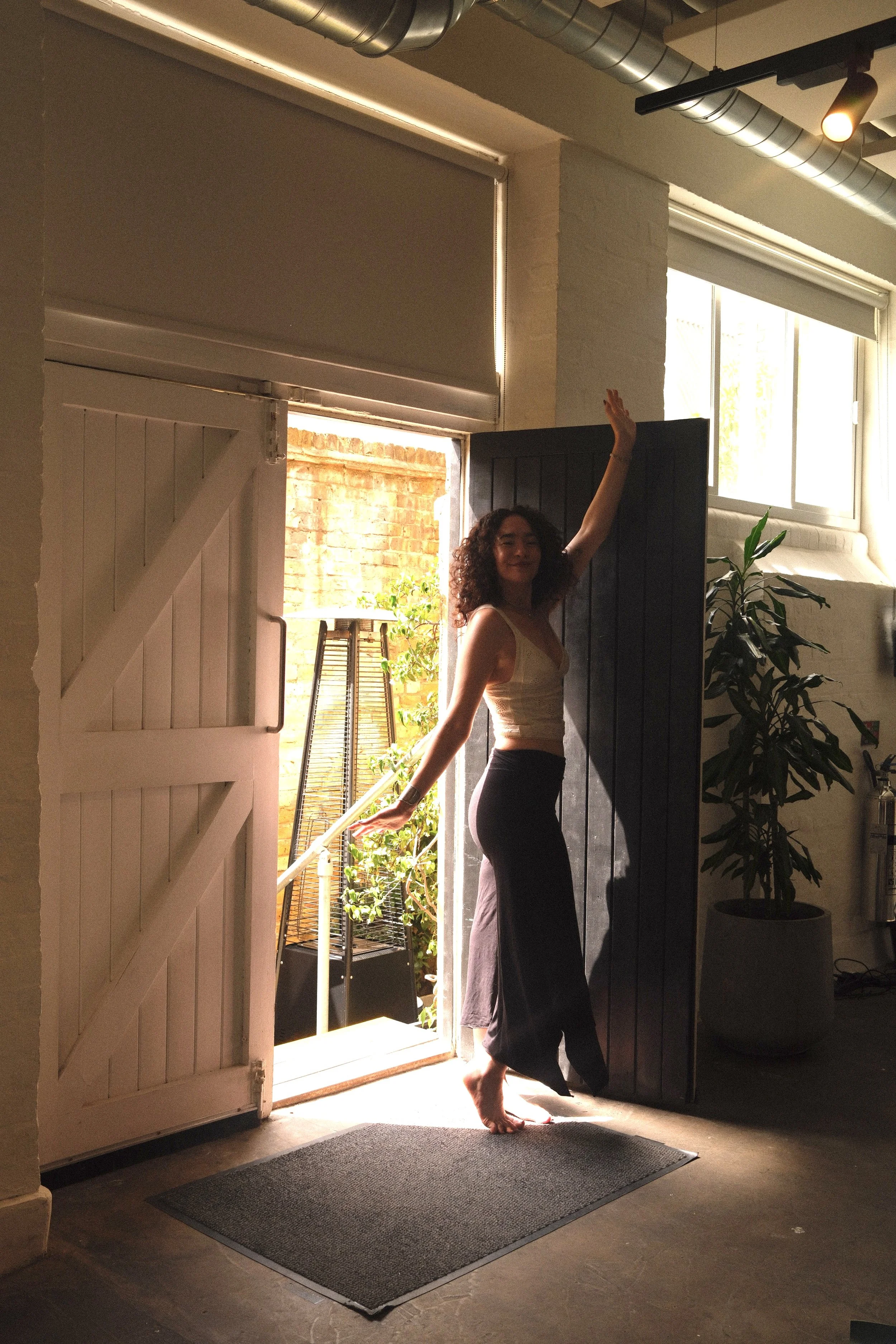 A woman with curly hair wearing a beige tank top and black pants stands barefoot in a doorway, smiling and raising one hand, inside a room with a greenhouse and plants visible outside.