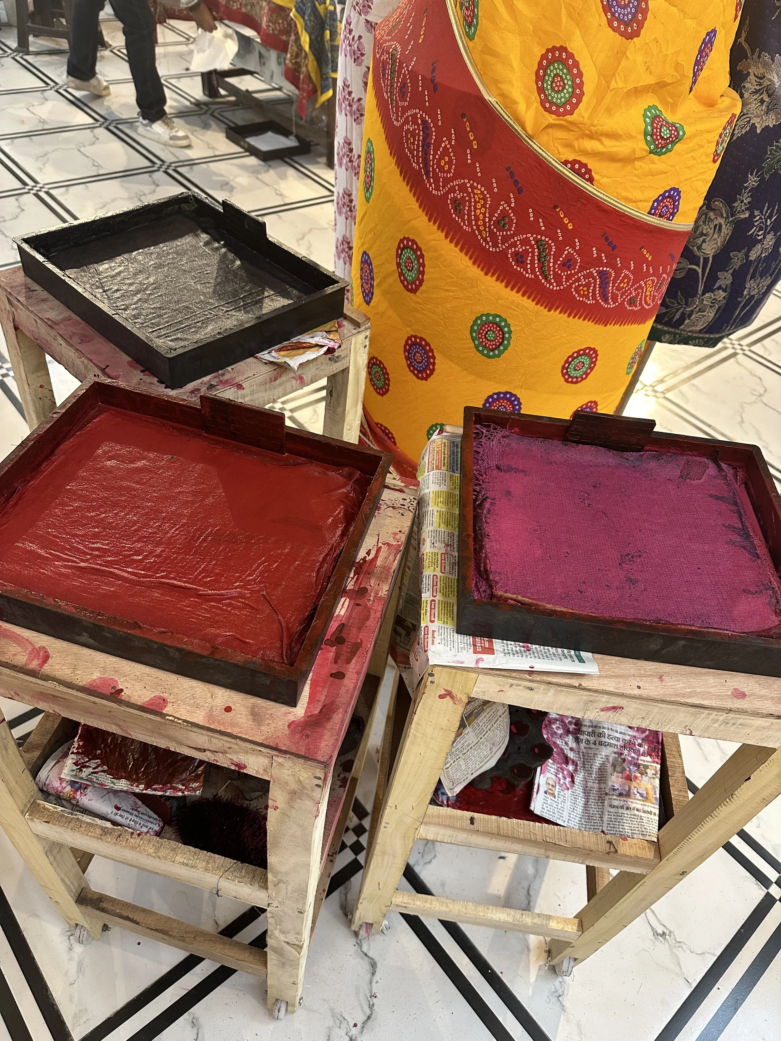 Three wooden trays with colored powders, black, red, and pink, set on small wooden tables lined with newspapers, in a marketplace or shop setting.  Meala Soul India retreat jaipur yoga retreat