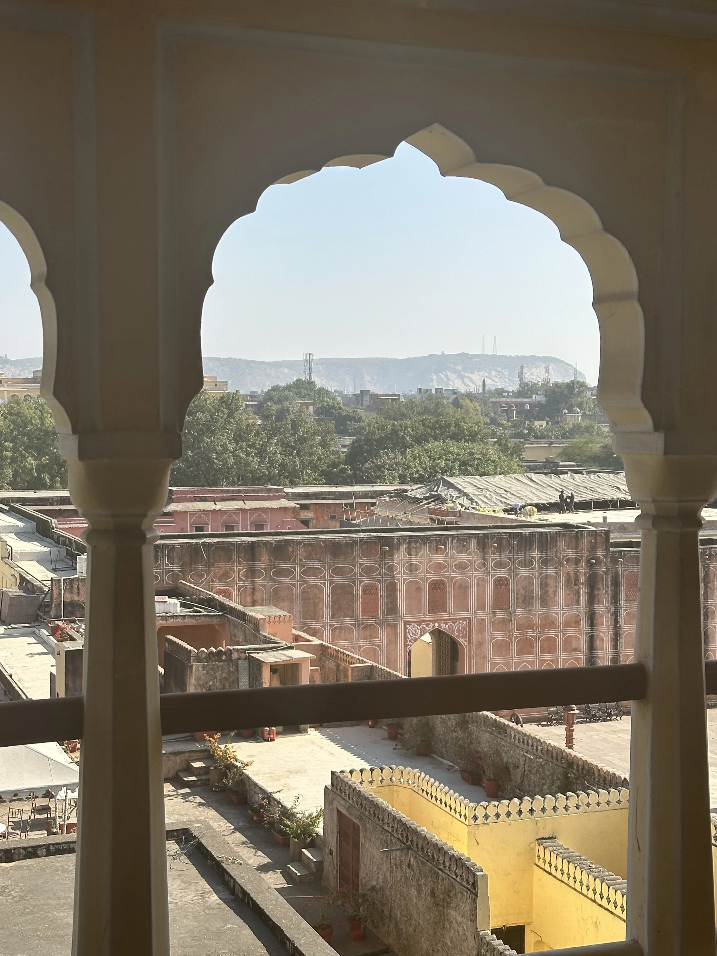   Meala Soul India retreat jaipur yoga retreat. View of rooftops and greenery through a decorative window in an ancient Indian palace or fort, with hills distant in the background.