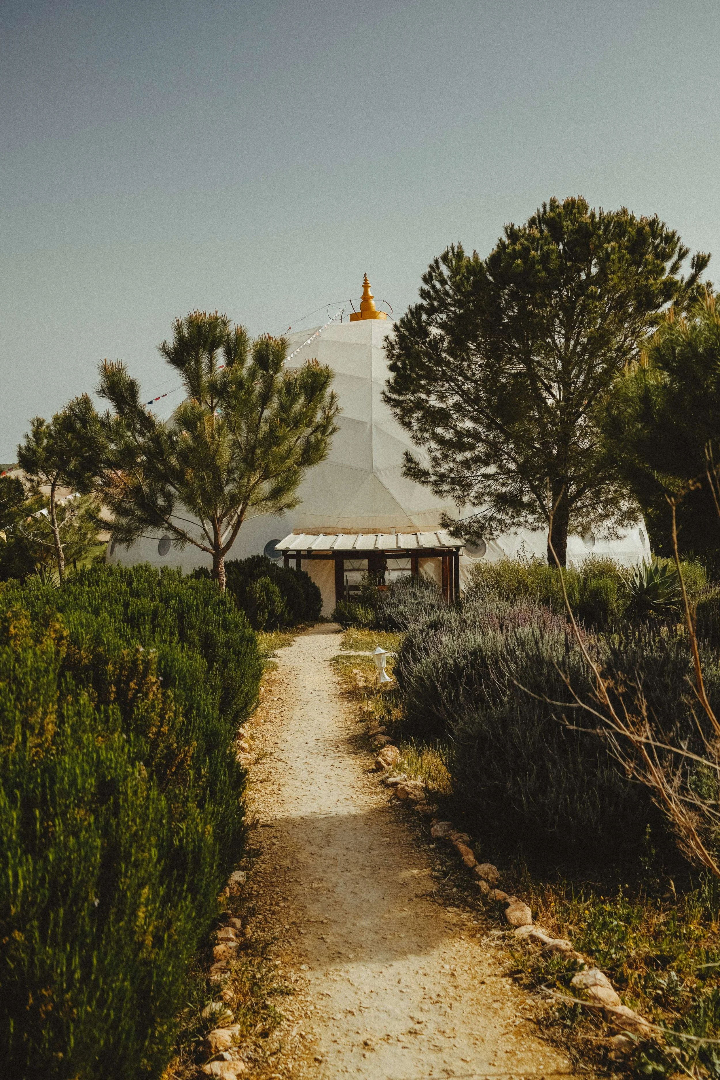 A dirt path leading to a white pagoda-like structure surrounded by trees and bushes under a clear sky. Meala Soul Yoga wellness retreat spain india womens retreat