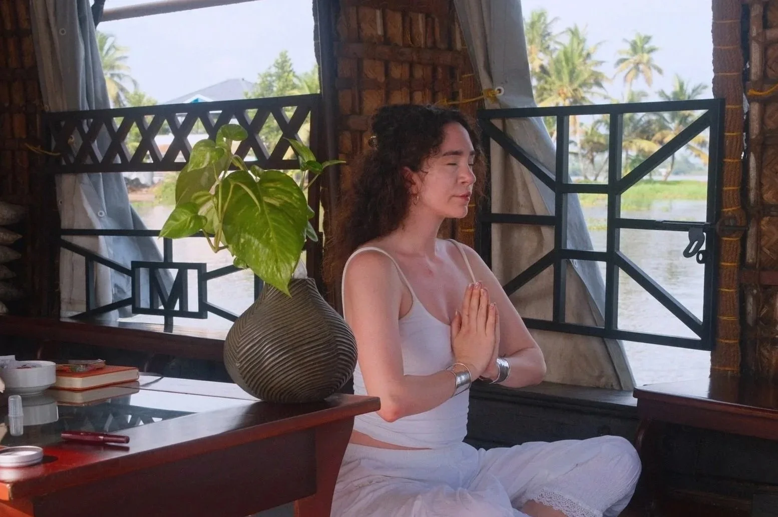 A woman practicing meditation indoors near a window with a view of palm trees and water, sitting cross-legged with hands in prayer position and eyes closed.