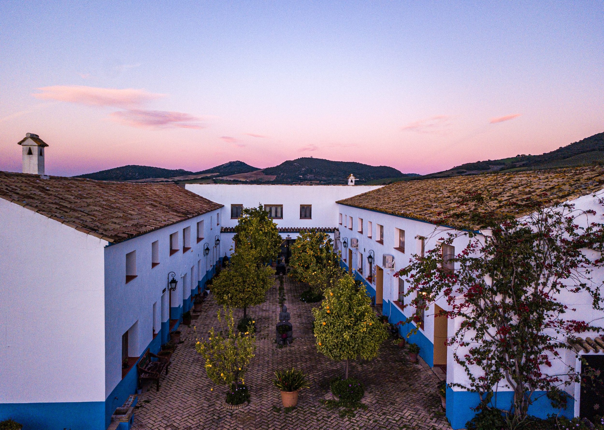 Yoga retreat Spain March 2027 Meala SoulViews of white buildings with brown tile roofs, a courtyard lined with trees and potted plants, and distant hills under a pink and purple sunset sky.