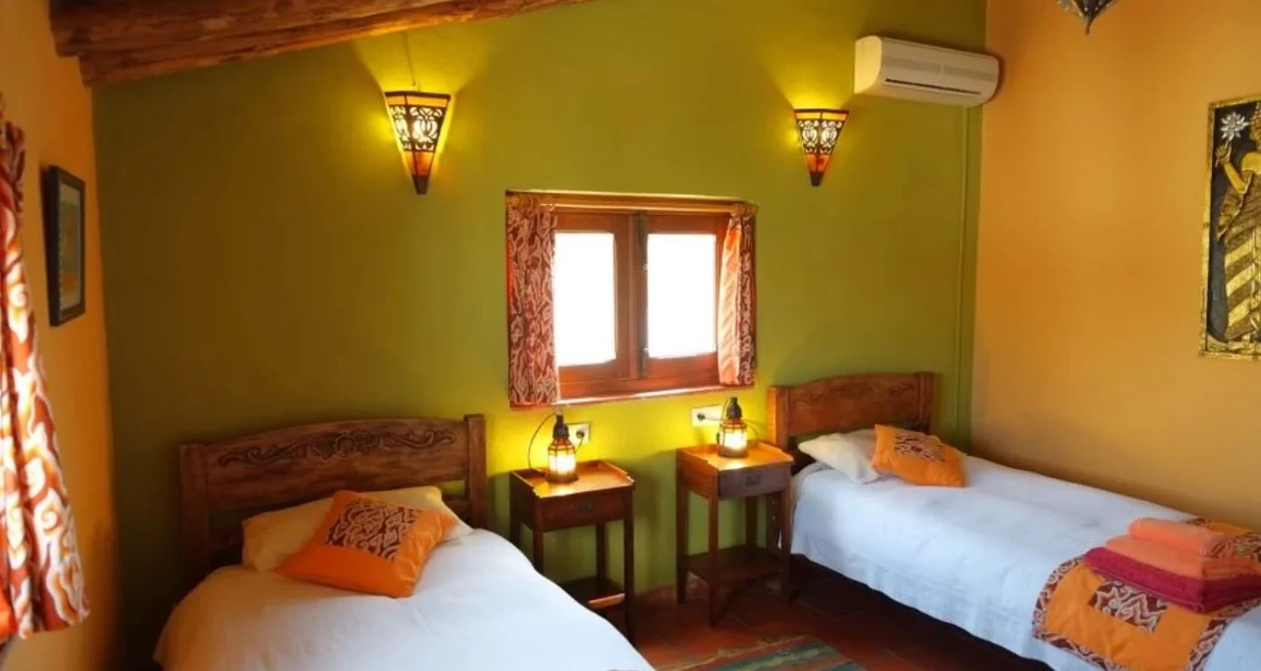 Two twin beds with wooden headboards in a colorful room with green and yellow walls. Between them are two small wooden nightstands with lantern-style lamps. A window with patterned curtains is centered between the beds. There is art on the walls, and a folded towel with a cloth is on one of the beds. An air conditioning unit is mounted high on the wall.