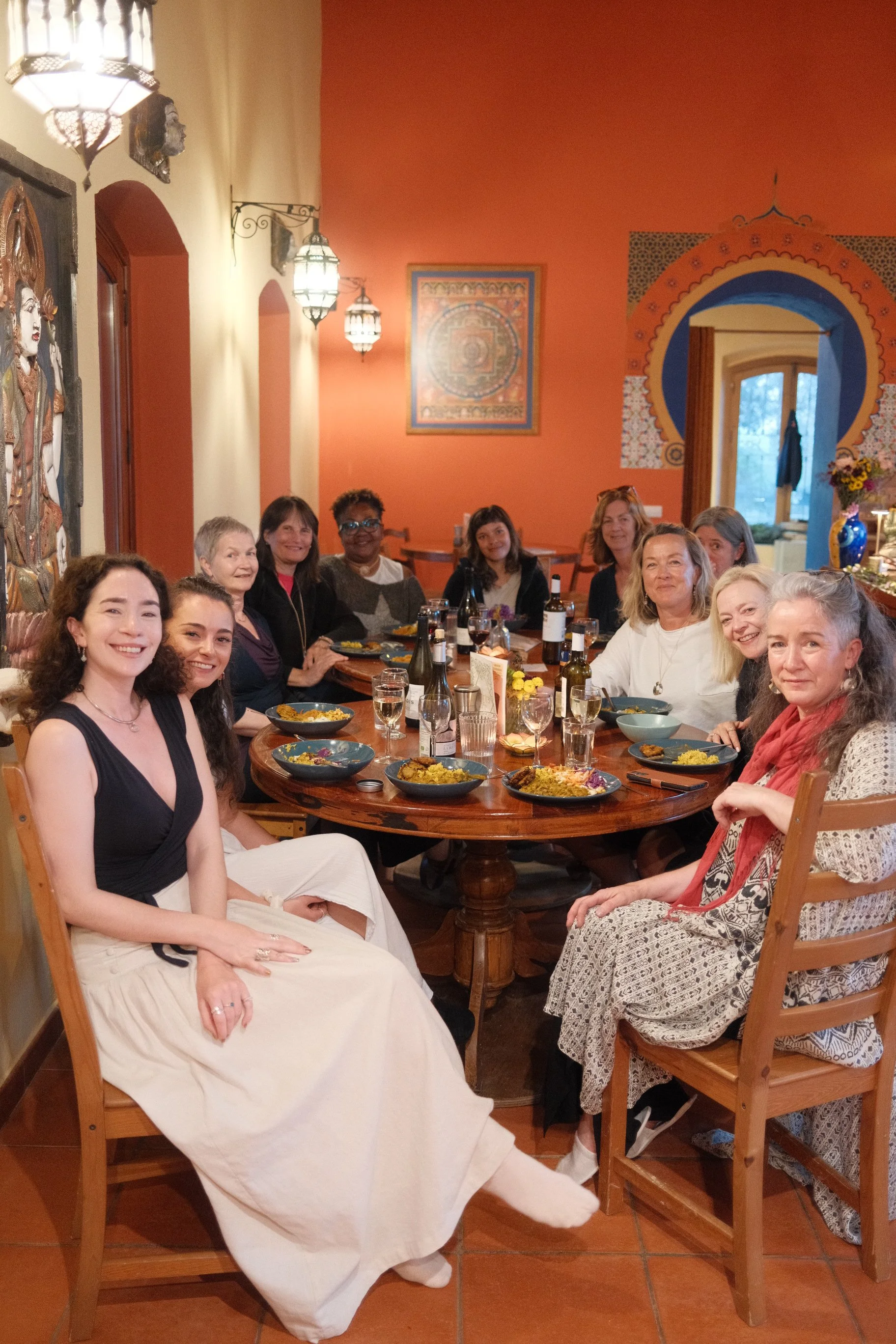 A group of women gathered around a dining table in a warmly decorated room with vibrant orange walls and traditional artwork, enjoying a meal together.