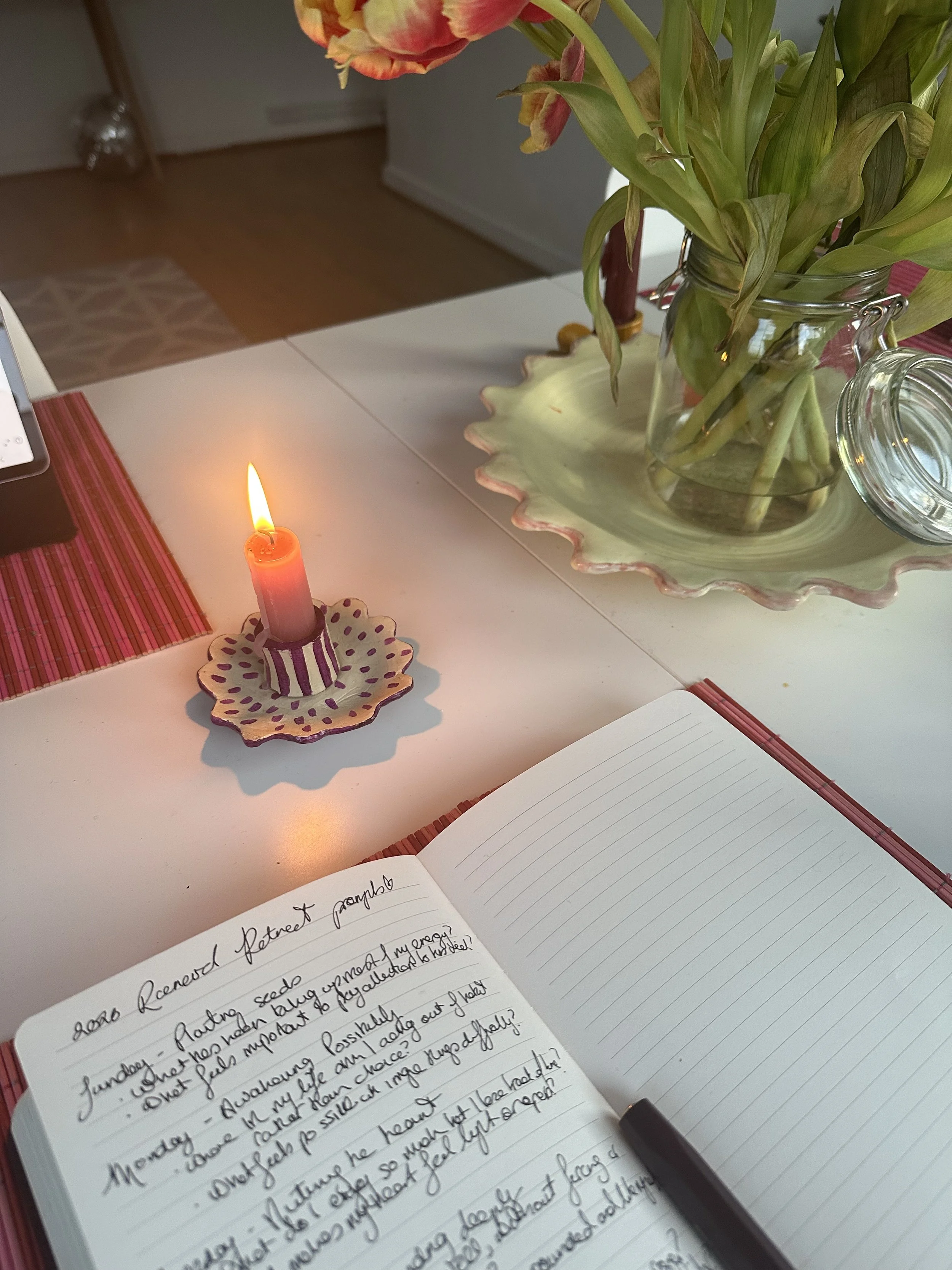 A white table with a lit pink candle in a decorative holder, an open notebook with handwritten notes, and a large glass vase with tulips. Red placemats are partially visible.