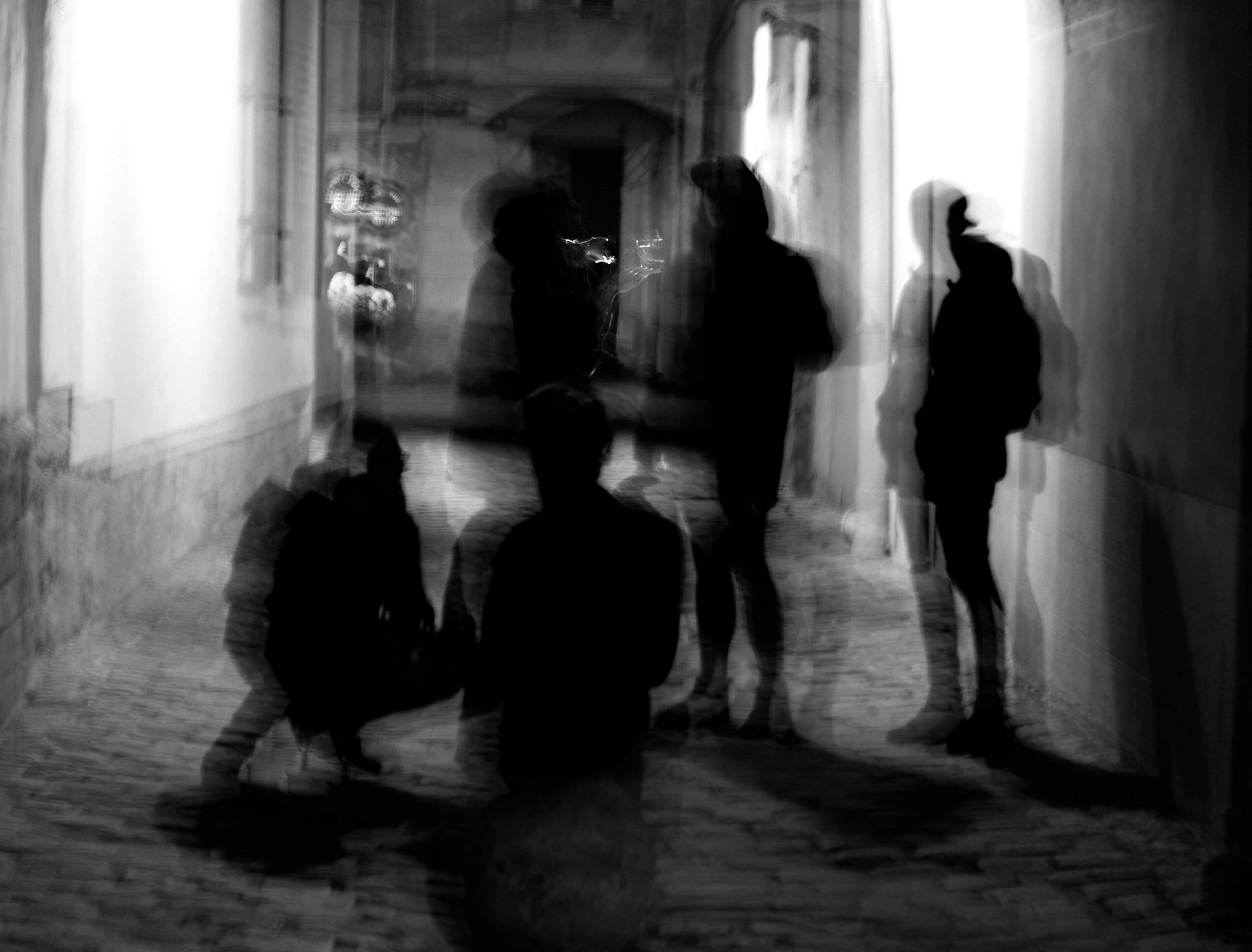 Black and white photo of five silhouetted people, some sitting and some standing, in a dimly lit hallway or corridor with an arched doorway in the background. Shadows and lighting create a layered, abstract effect.