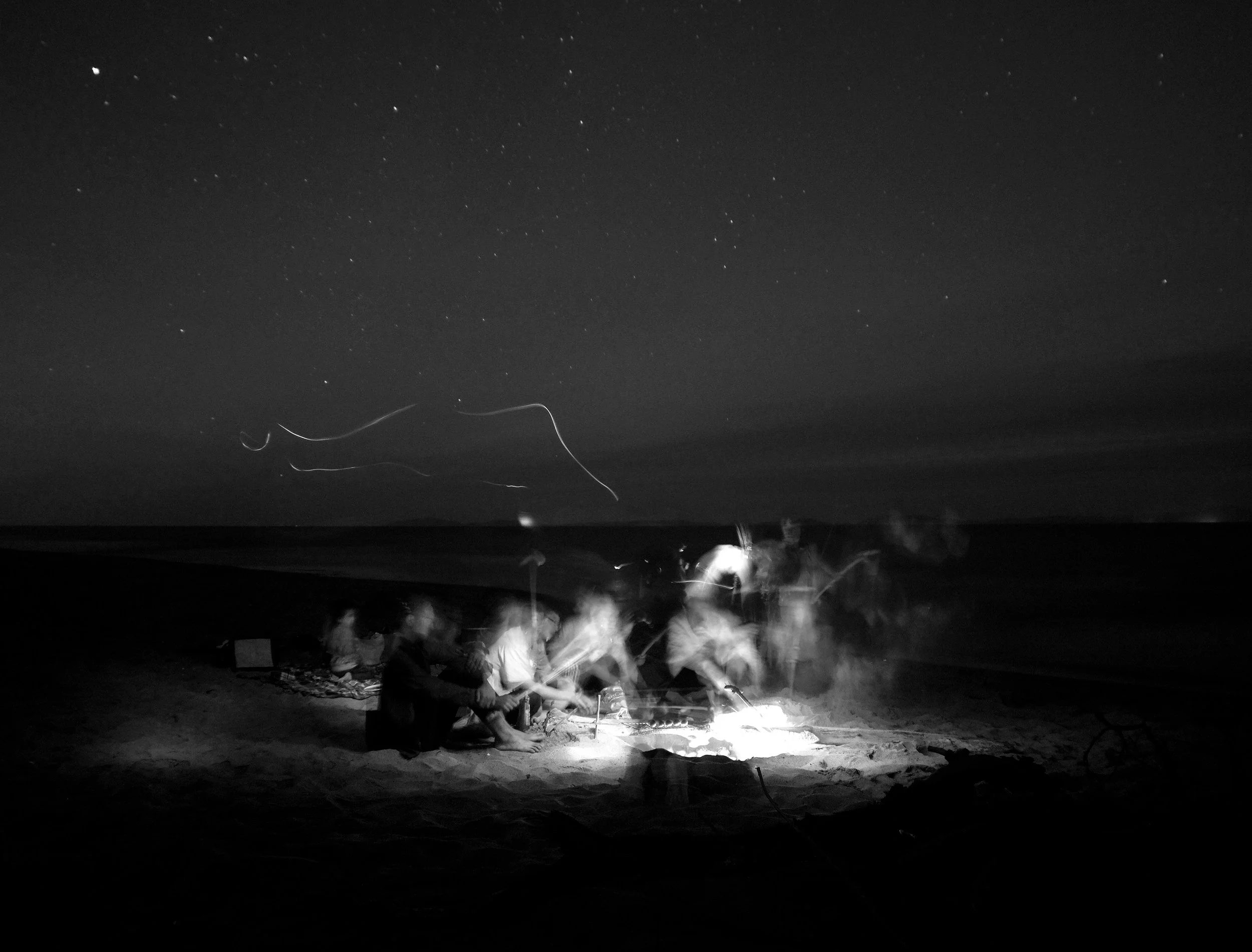 People sitting around a beach campfire at night under a starry sky with light trails in the air.
