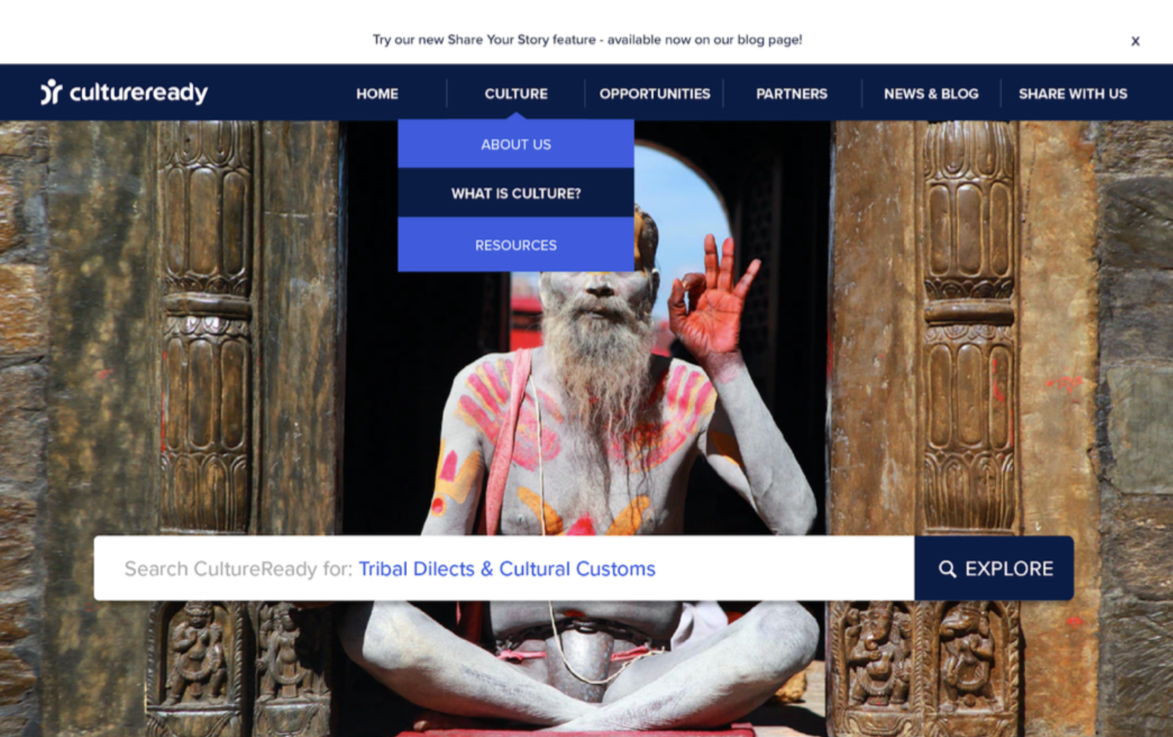 A person painted white, sitting cross-legged in front of a carved wooden frame, performing a gesture with their hand, at a cultural site with stone walls.