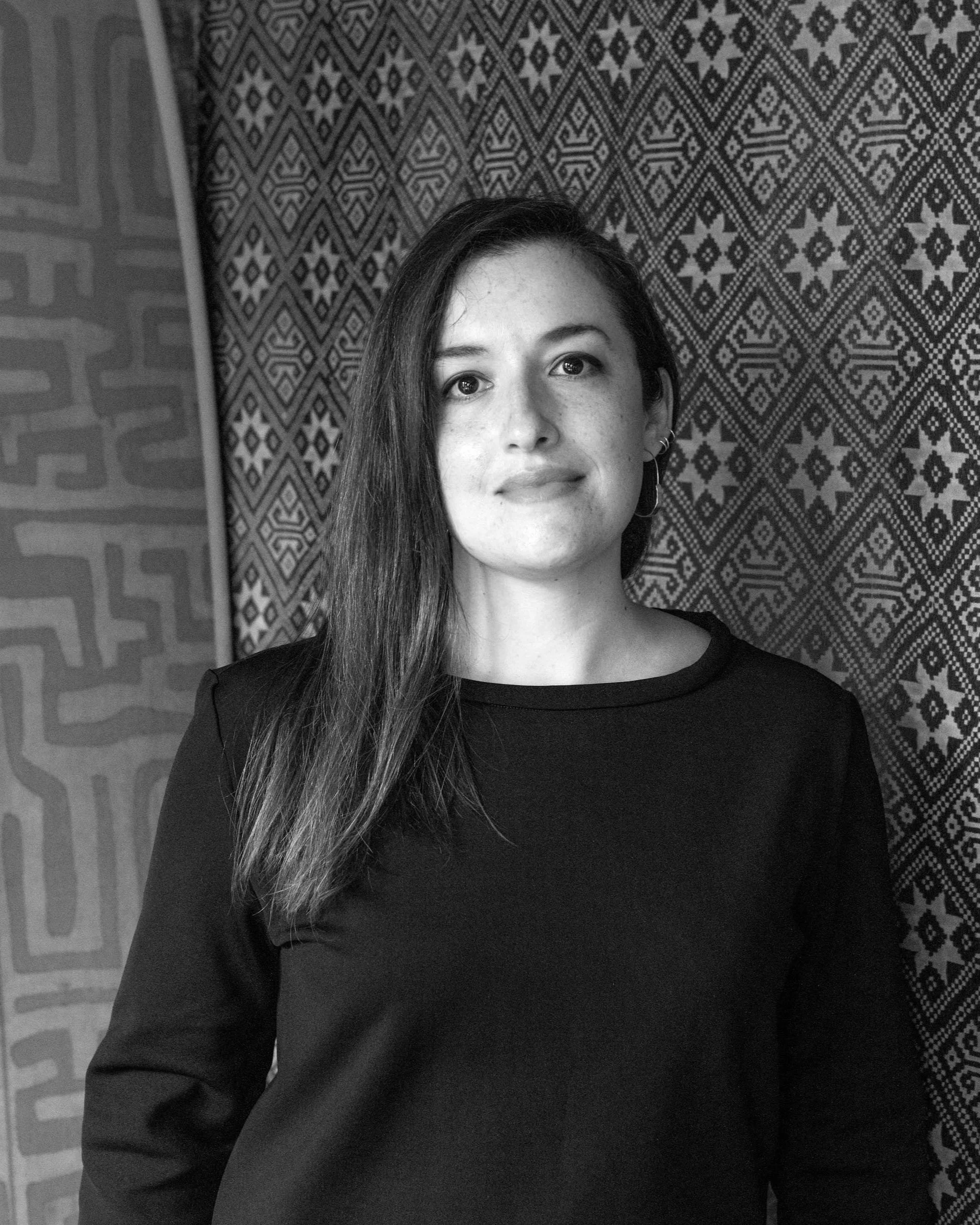 A woman with long dark hair and earrings, wearing a black shirt, standing in front of patterned fabrics.