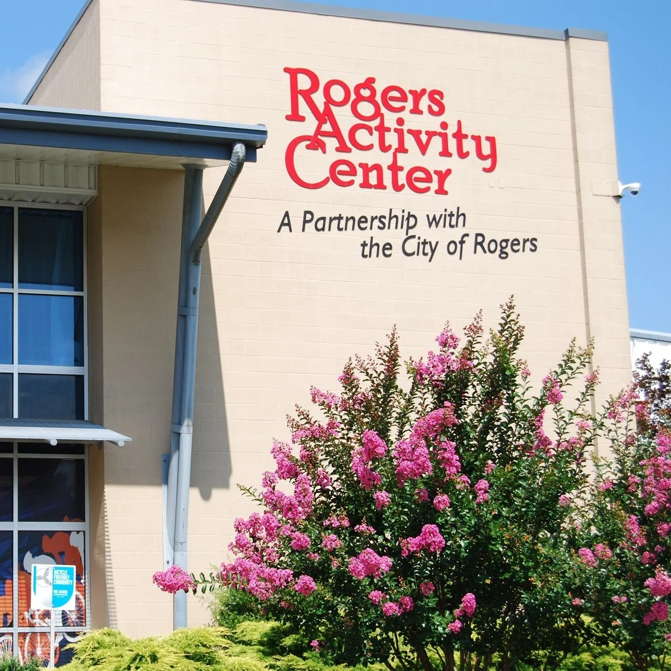 Rogers Activity Center building facade with sign, partnership mention, and blooming pink flowers.