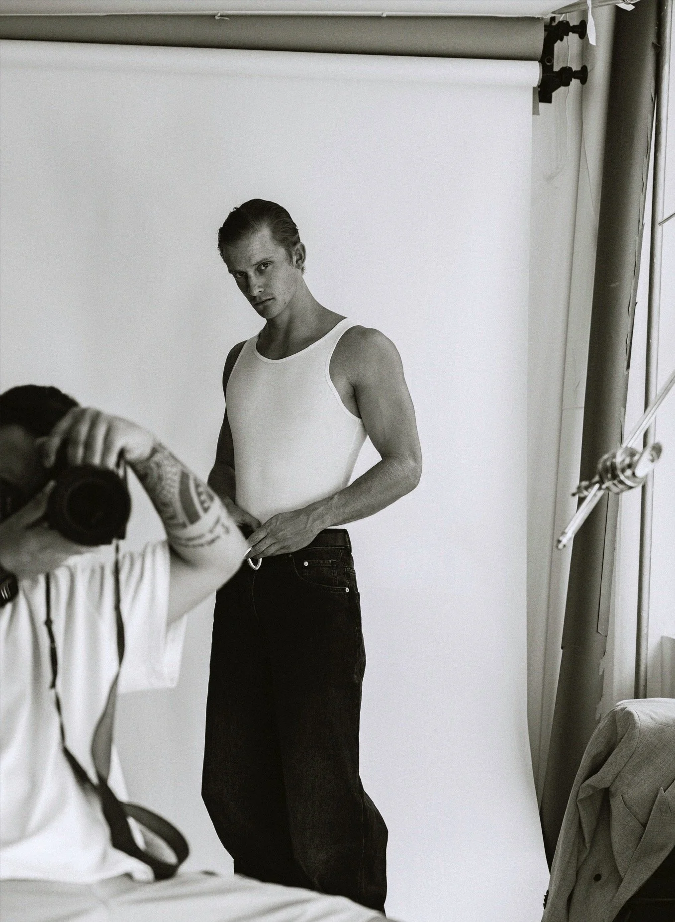 Black-and-white photo of a young man with slicked-back hair wearing a sleeveless white tank top and dark jeans, standing against a plain backdrop during a photo shoot.