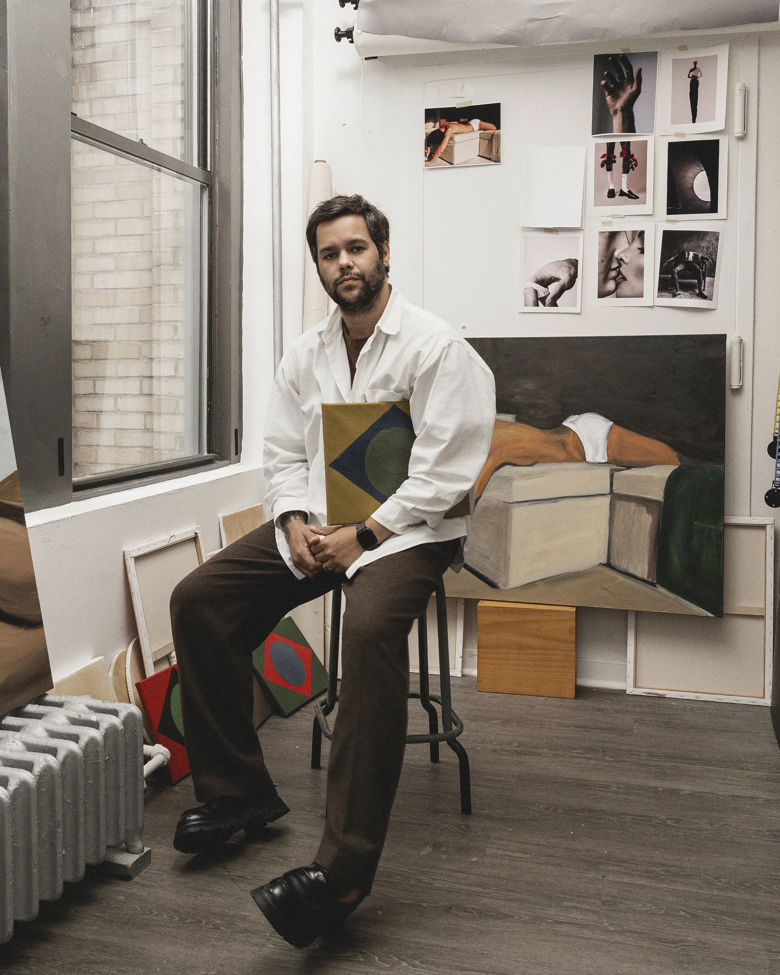 A man with dark hair and a beard sitting on a stool in an art studio, holding a piece of art. The studio has several paintings and photographs on the walls and leaning against objects, with a large window. Self portrait of Ton Gomes