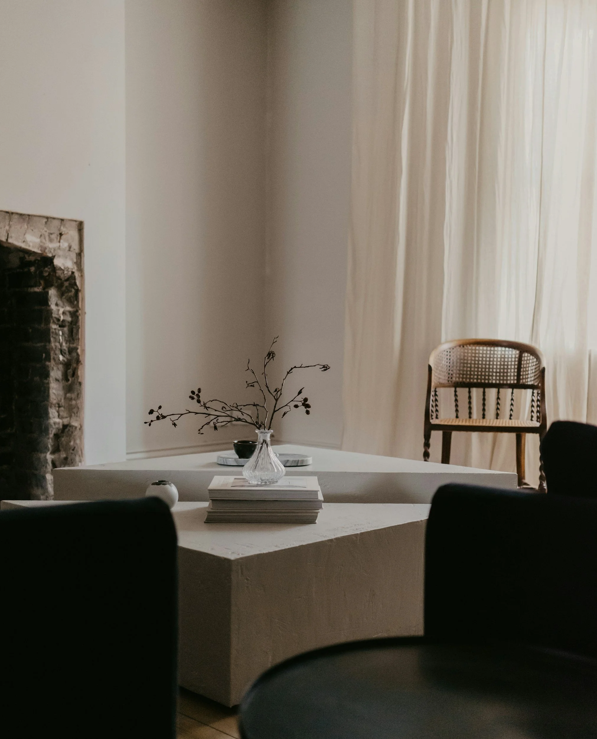 Minimalist living room with a geometric table, decorative vase with branches, stack of books, wicker chair, sheer curtains, and exposed brick wall.