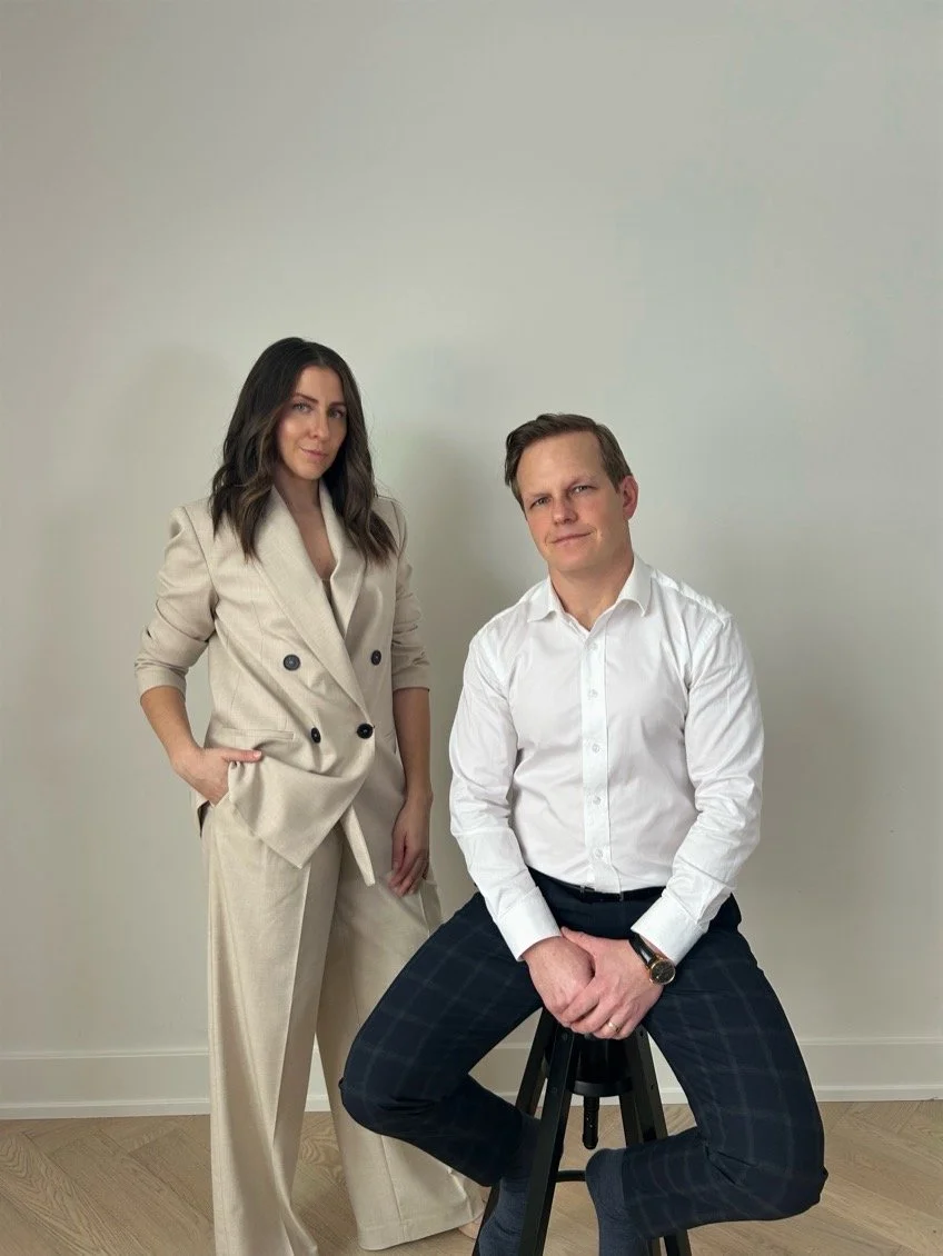 Two people posing; a woman in a beige suit standing and a man in a white shirt seated on a stool, against a plain background.