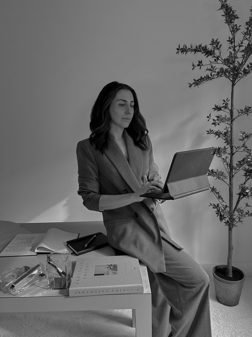 Woman in suit working on a laptop, seated on a table with notebooks, a pen, and a book titled 'Arranging Things,' next to a potted plant.