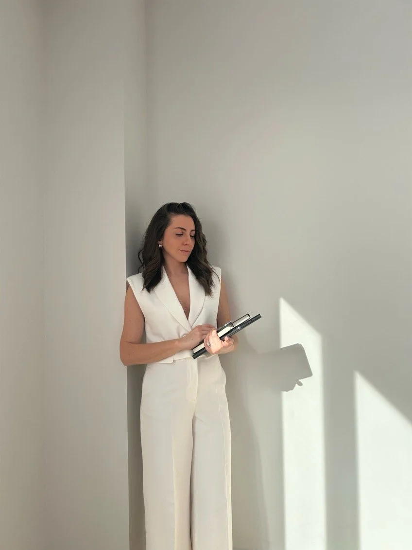A woman in a white sleeveless jumpsuit stands against a wall holding a notebook. The scene is minimalistic with soft, natural lighting.