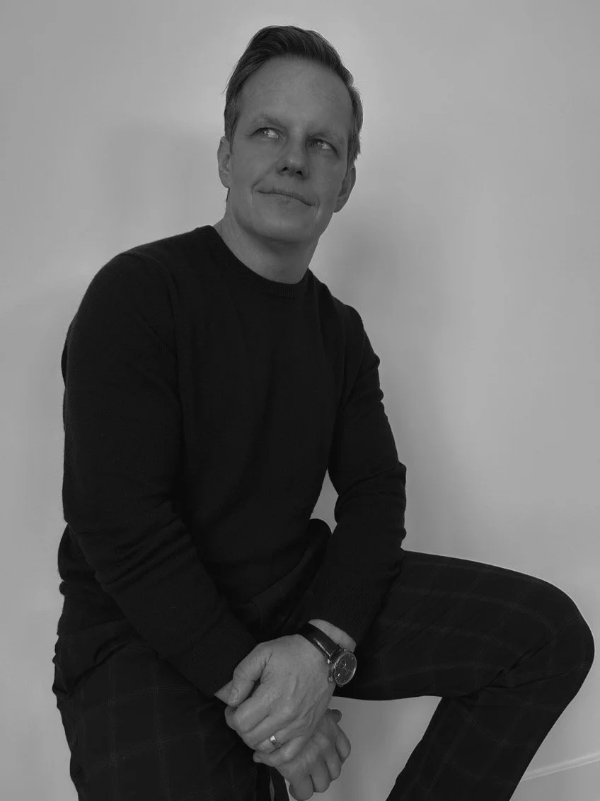 Black and white photo of a person wearing a dark long-sleeve shirt, sitting with one hand resting on their knee.