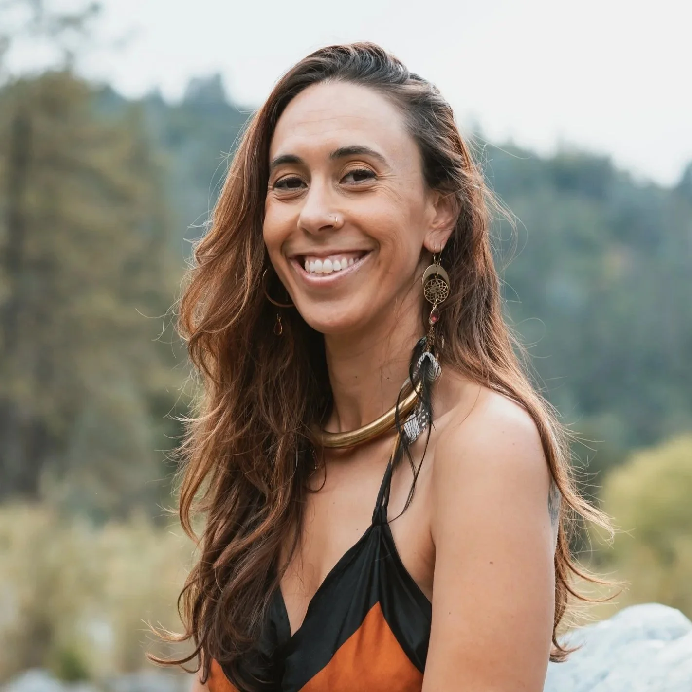 A woman with long, wavy brown hair smiling outdoors in a natural setting, wearing earrings and a sleeveless top.