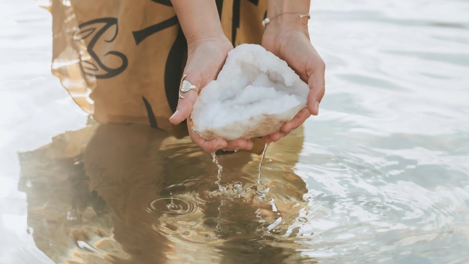 A person is standing in shallow water, holding a large white crystalline geode dripping water with both hands.