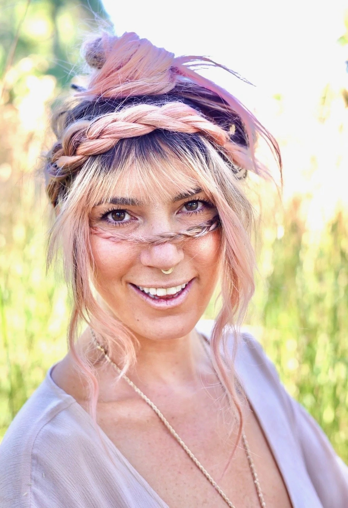A woman with pink hair styled with braids and loose waves, smiling outdoors in sunlight.