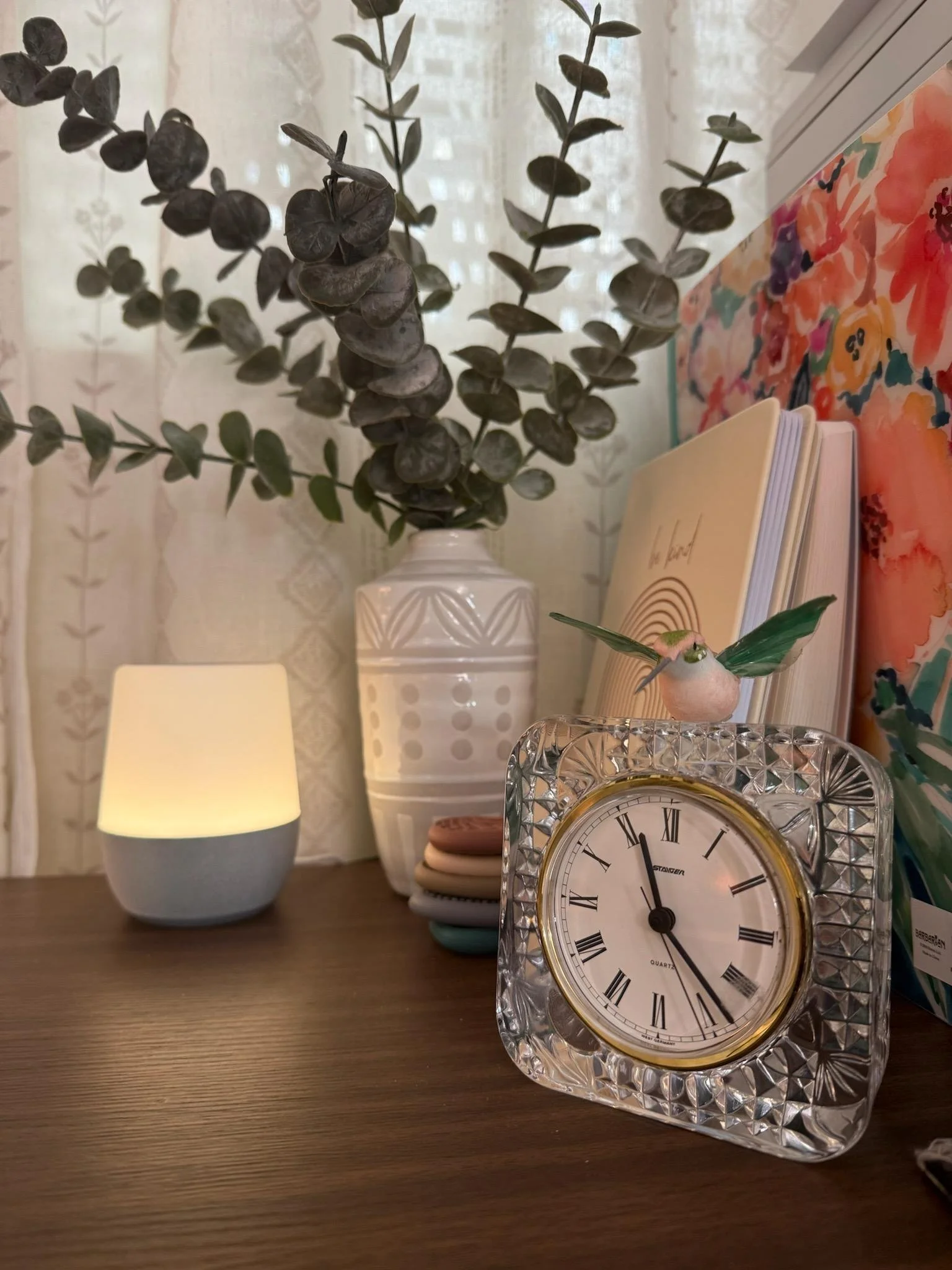 Decorative tabletop scene with a vase of eucalyptus, a small lamp, a stack of notebooks or coasters, a bird figurine with green wings, a vintage-style clock, and colorful artwork in the background.