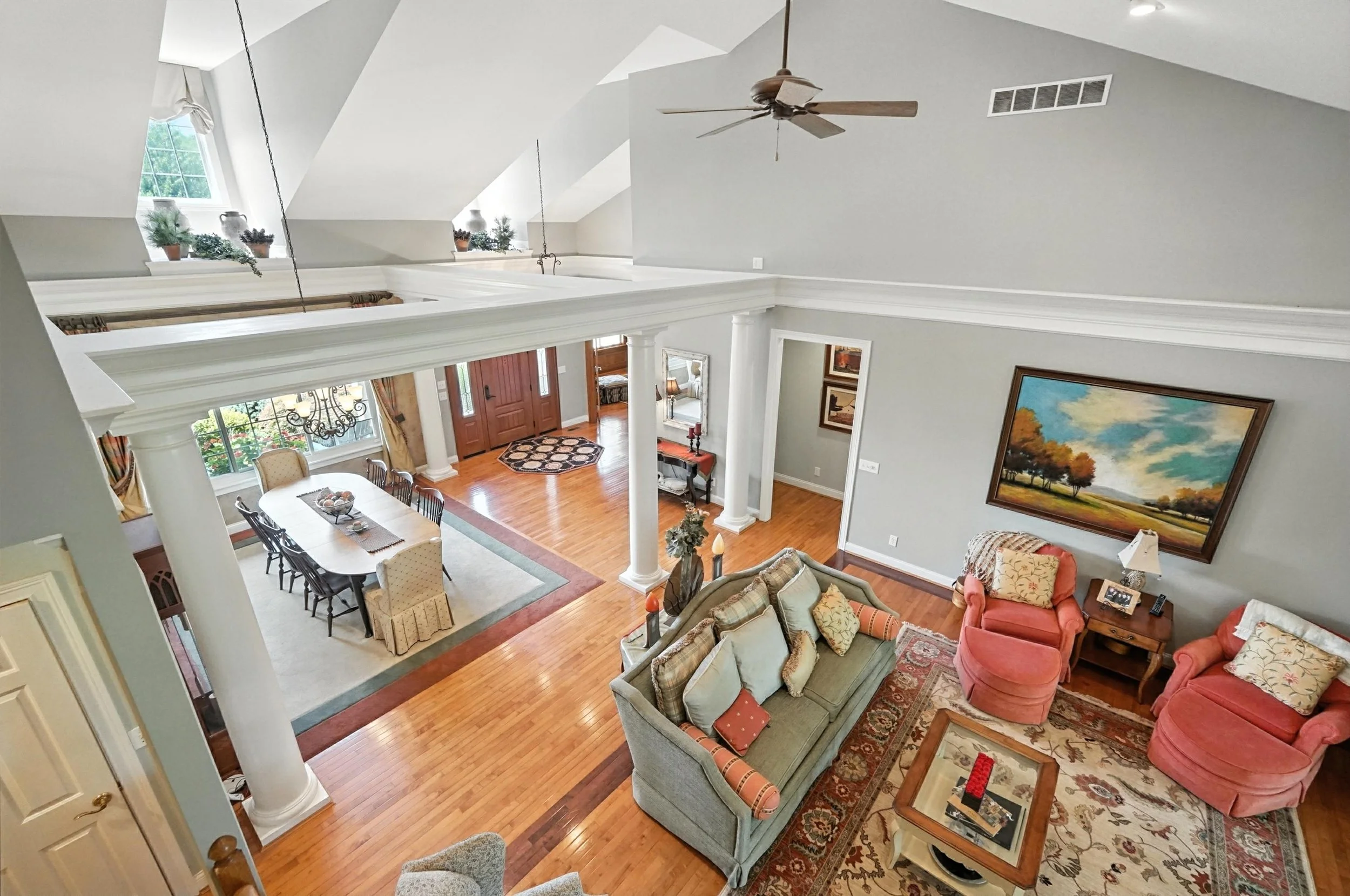 Living and dining room with hardwood floors, light gray walls, white columns, large windows, and a ceiling fan. The living room has a sofa, two pink armchairs, a coffee table, and wall art. The dining area features a large table with eight chairs, a