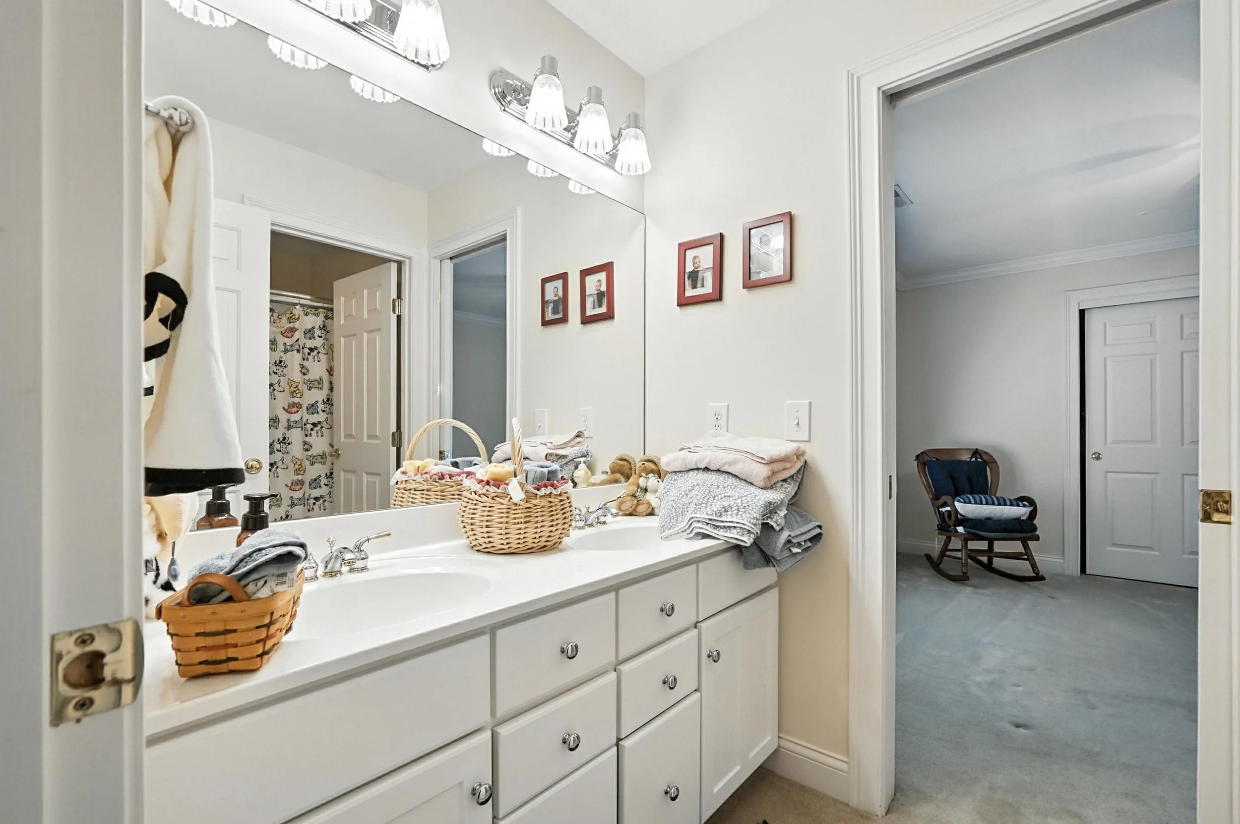 Bathroom vanity with cluttered towels, baskets, and toiletries, large mirror, framed pictures on the wall, doorway to a room with a rocking chair and closet.