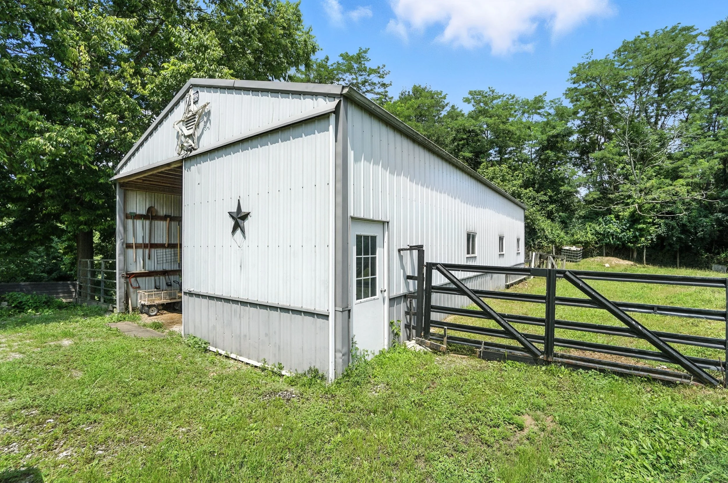White metal barn with star decorations, surrounded by green grass and trees, with a black gate and a small door, under a blue sky with clouds.