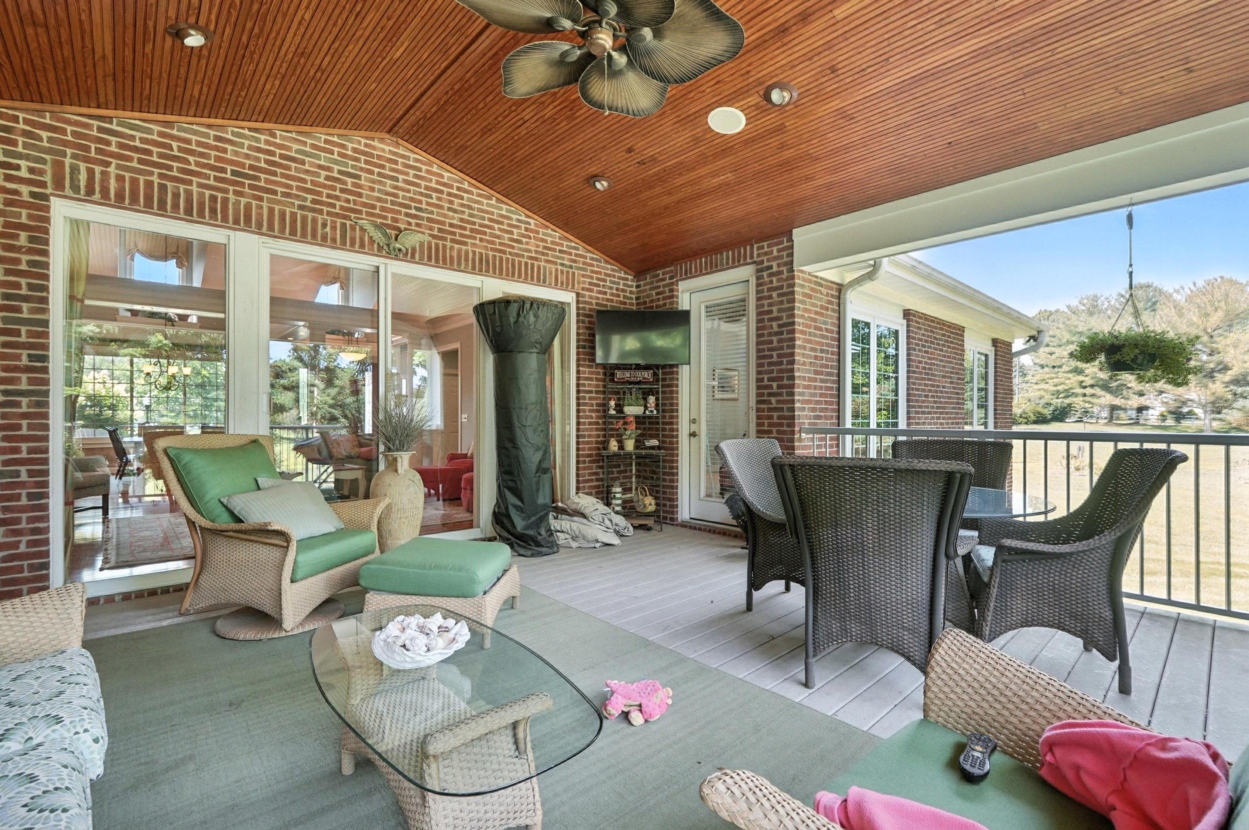 Covered outdoor patio with wicker chairs, a glass-topped table, potted plants, a ceiling fan, and a brick wall with sliding glass doors leading to an interior room.