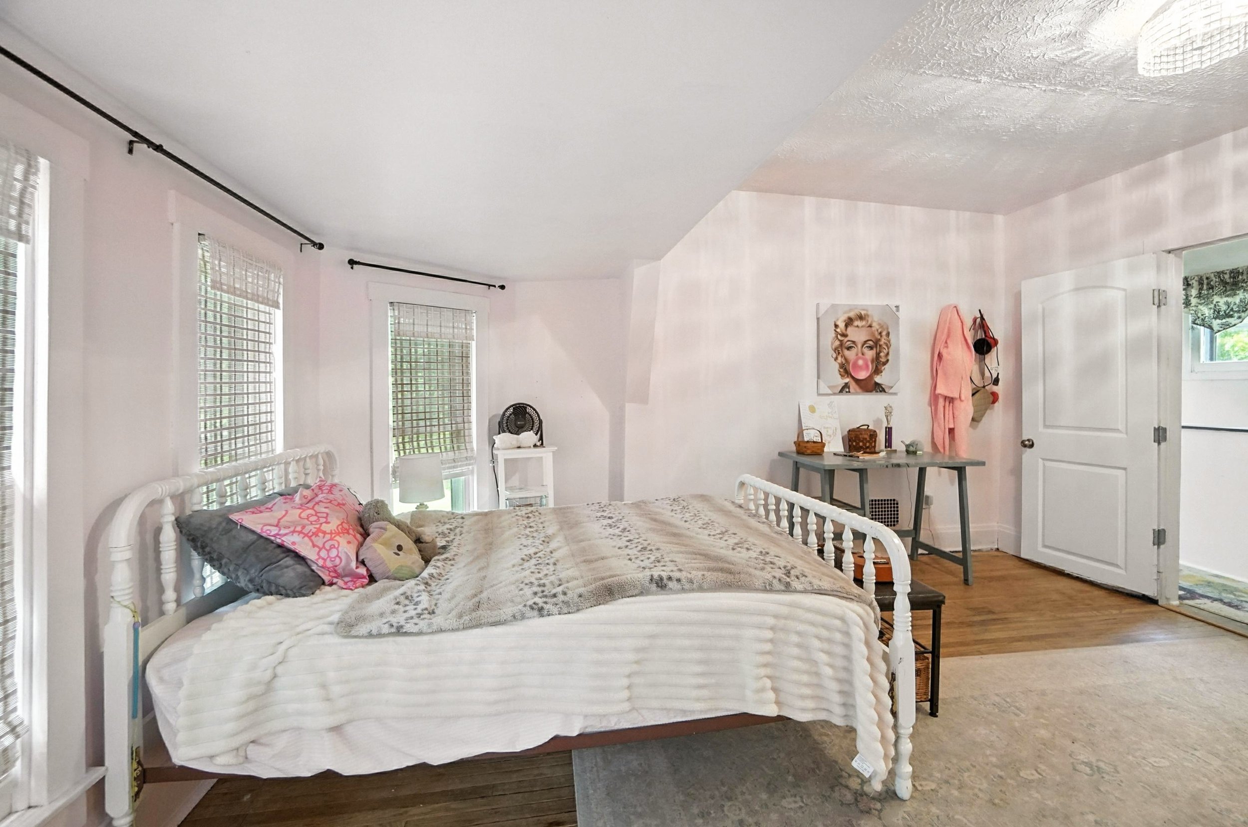 Bedroom with white bed frame, pink and gray pillows, beige blankets, white wall with windows, art on wall, gray dresser, door open to another room.
