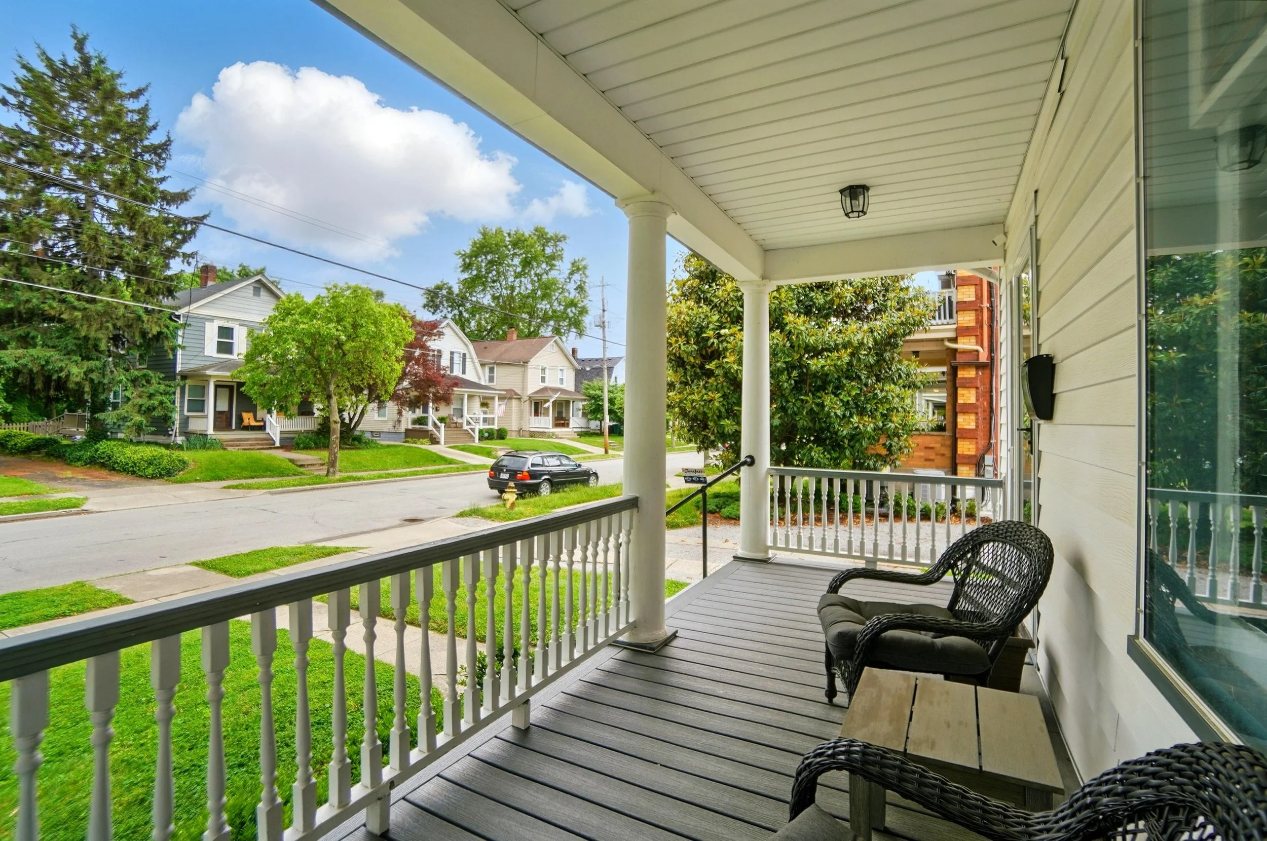 View from a house porch looking out over a residential street with trees and houses, with chairs and a small table on the porch.