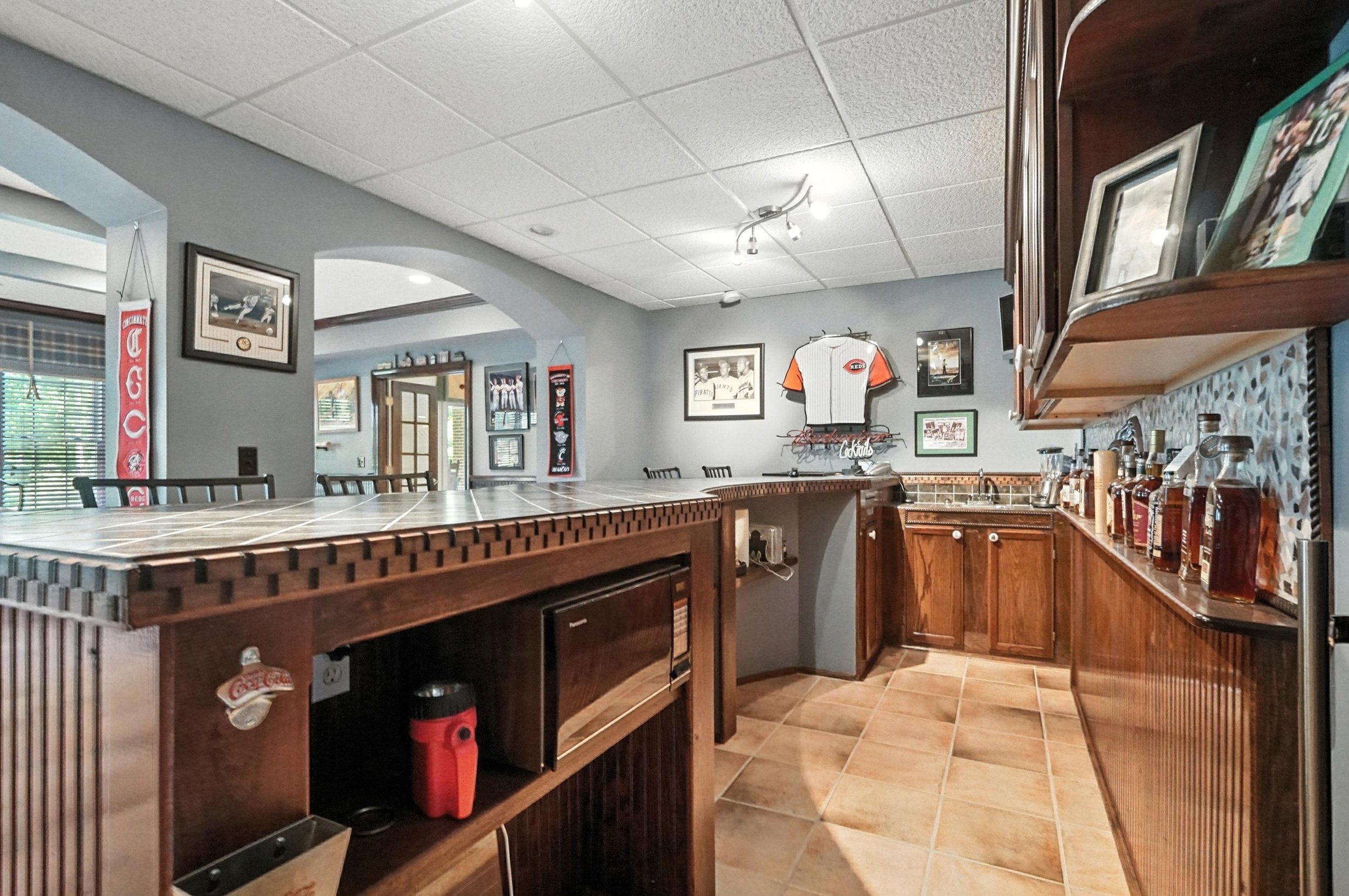 Home bar with wooden cabinets, tiled countertop, framed sports memorabilia on the wall, and a collection of liquor bottles on the counter.