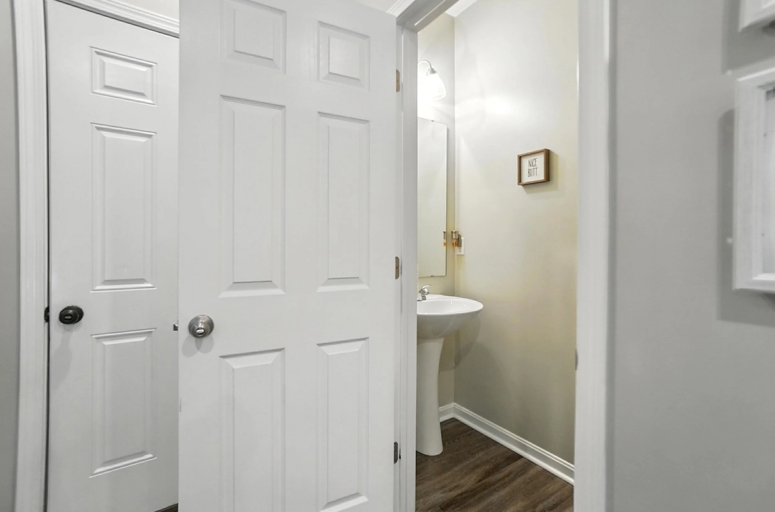 View of a small bathroom with a sink and mirror, partially seen through open white closet doors. A framed sign on the wall reads 'Nice Butt.' The bathroom has beige walls and dark wood flooring.