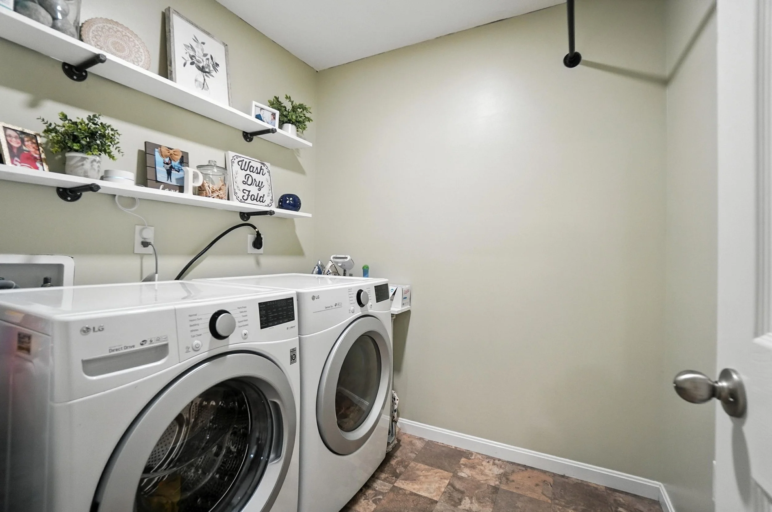 A laundry room with a front-loading washing machine and dryer, white shelves with decorations and laundry supplies, and a beige painted wall.