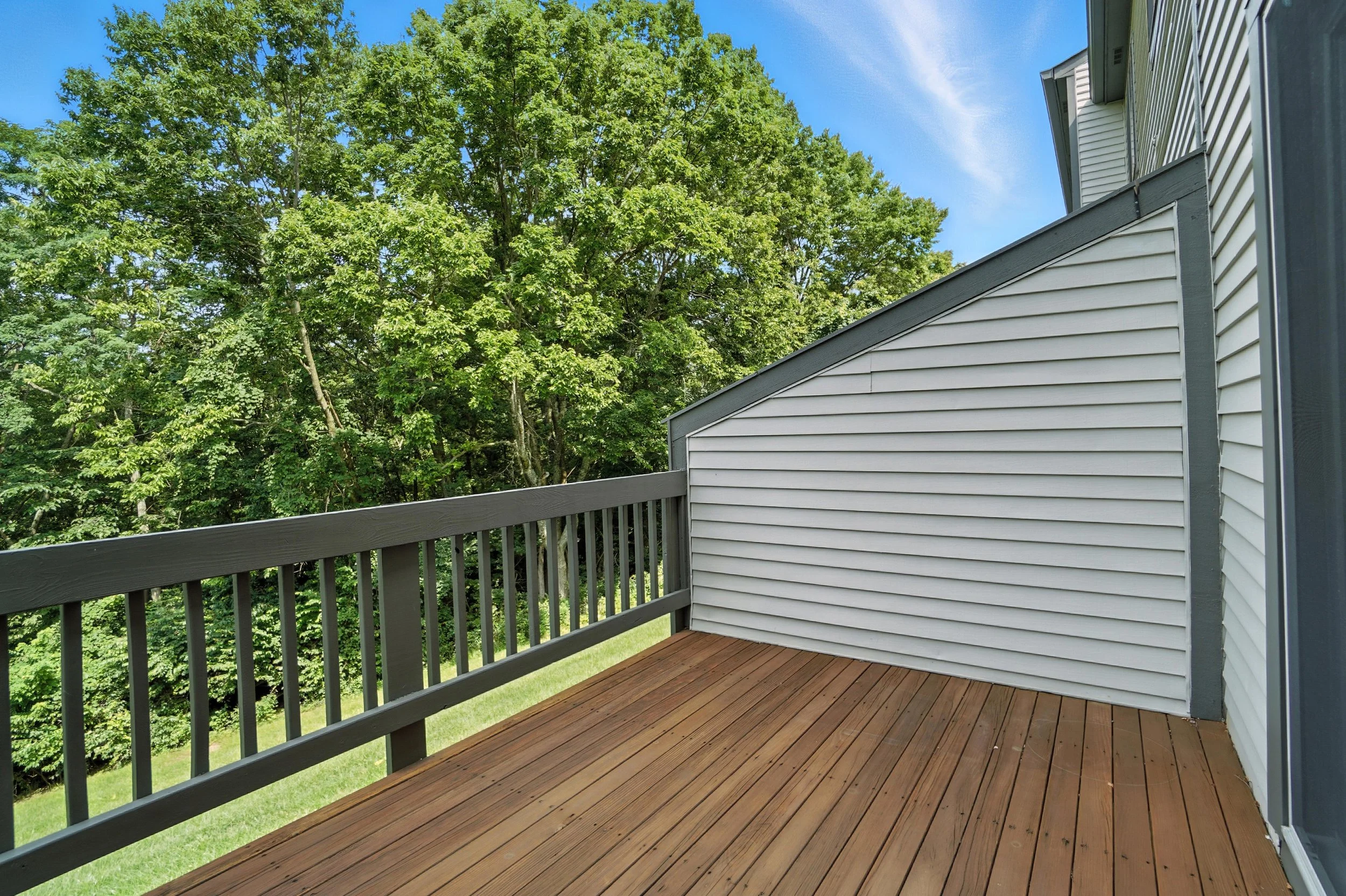 Empty wooden balcony with railing, adjacent to house with horizontal siding, overlooking lush green trees under a blue sky.
