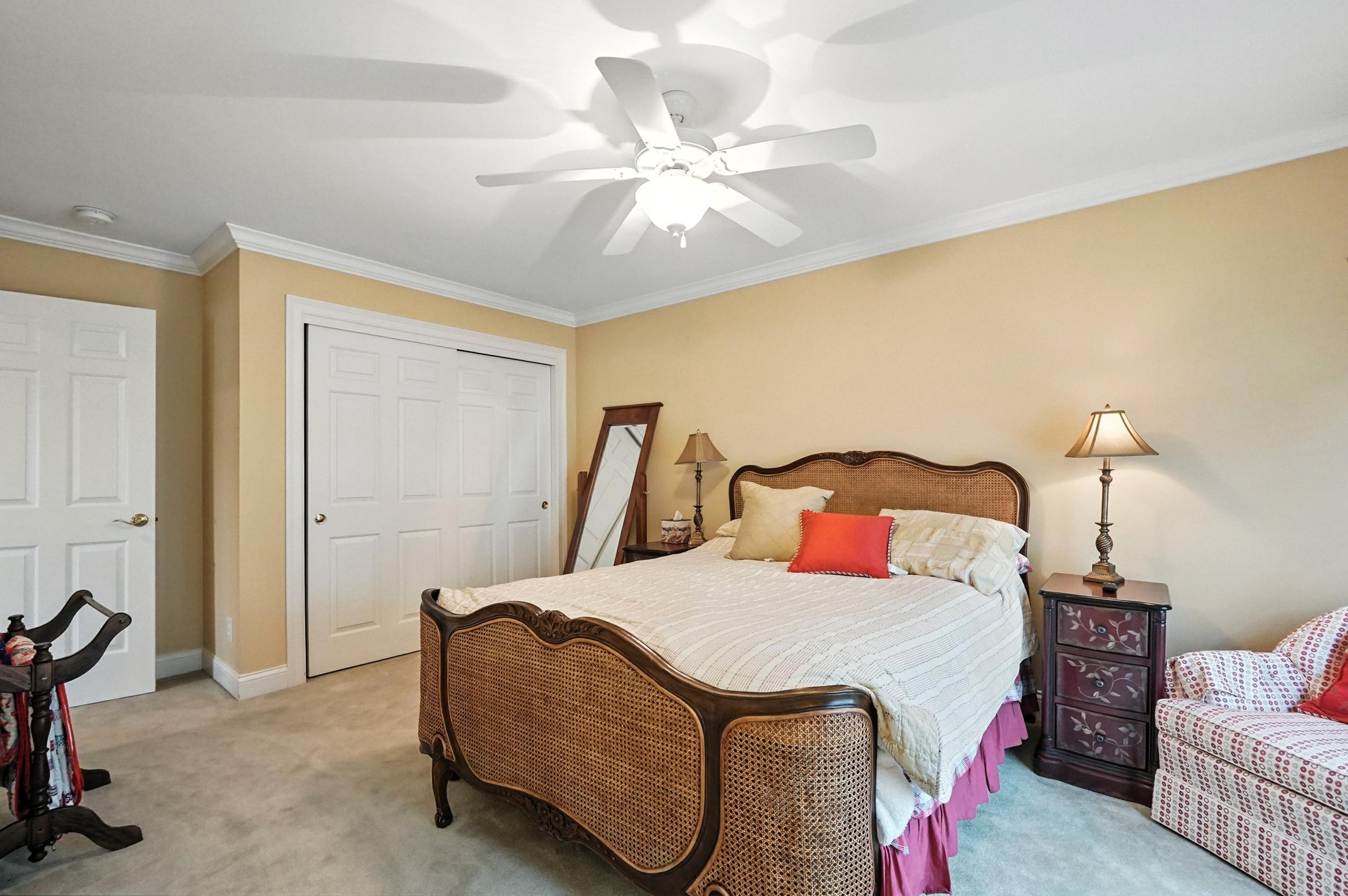 A bedroom with a wooden bed, white bedding, a red and beige pillow, a wooden nightstand with a lamp, and a wall mirror. There is a white ceiling fan and closet doors on the wall.