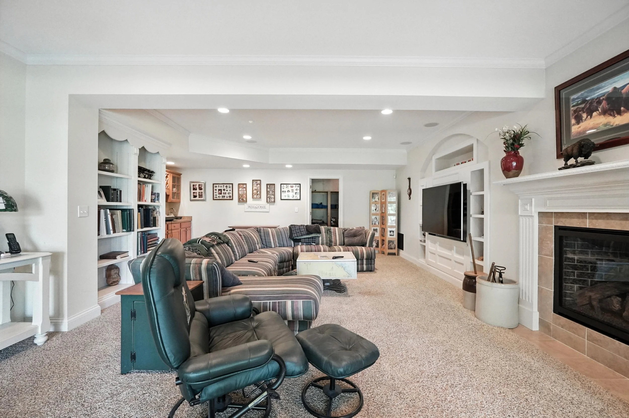 Living room with a patterned sofa, black leather recliner, built-in white entertainment center with TV, fireplace, and decorative items, carpeted floor, and framed pictures on the wall.