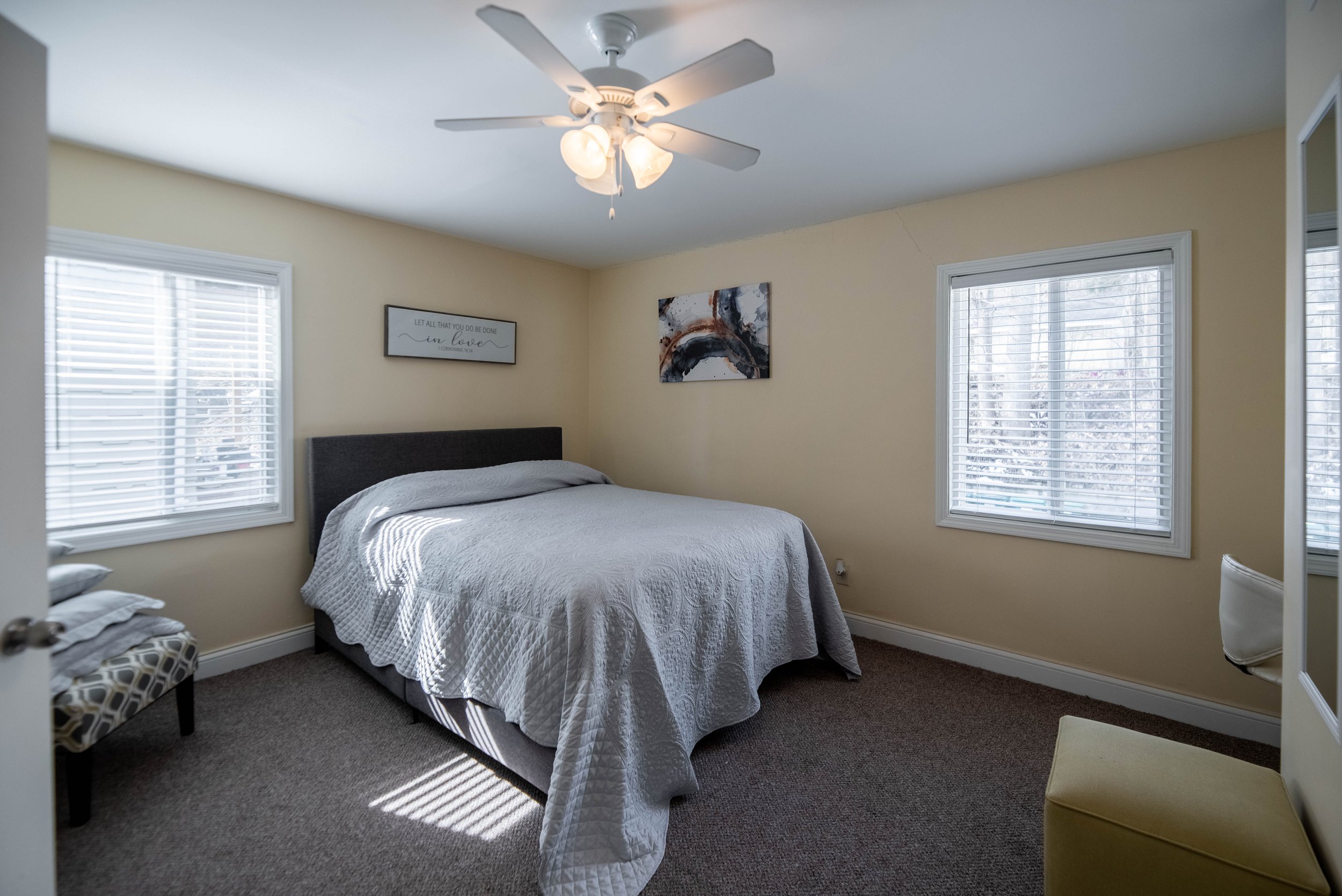 Cozy bedroom with a double bed, gray quilt, ceiling fan, two windows with blinds, wall art, and a small bench with cushions.