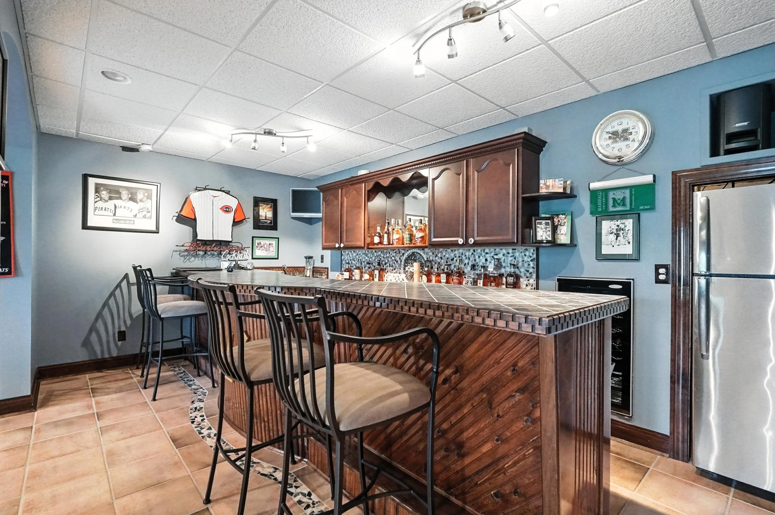 Home bar with wooden cabinets, tiled counter, bar stools, and various framed sports memorabilia on blue walls.