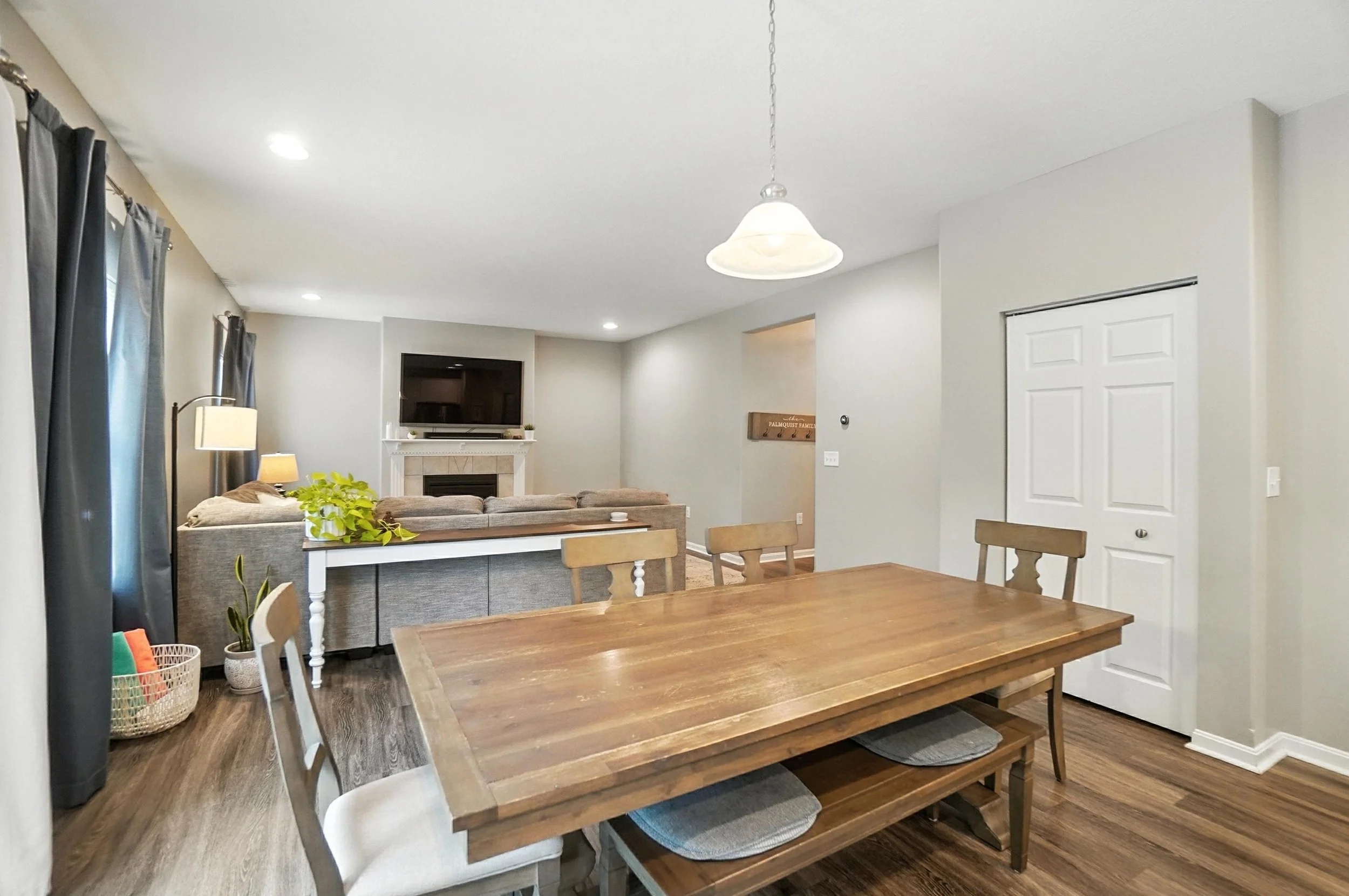 Open-concept living and dining area with a wooden dining table, beige chairs, a gray sofa, a fireplace with a mounted flat-screen TV, and large windows with dark curtains.