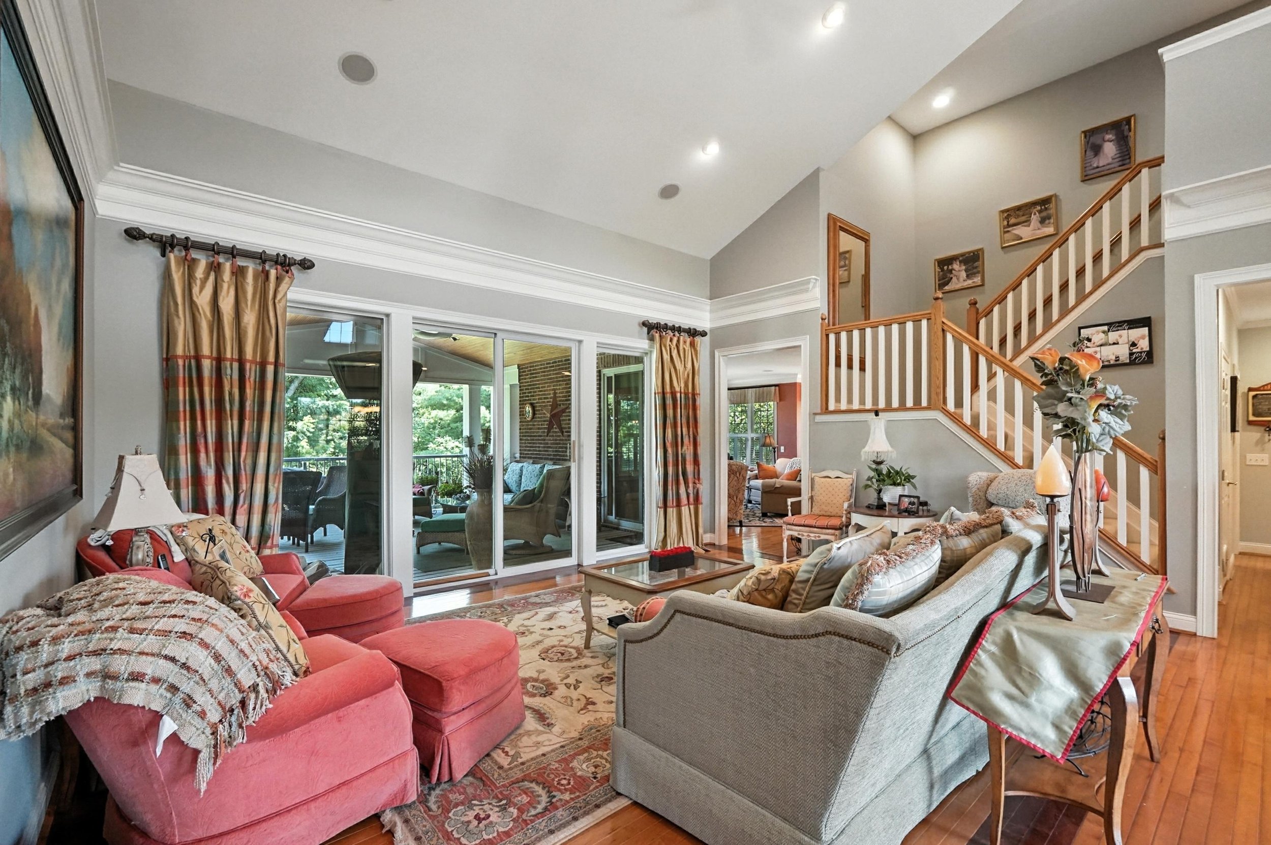 Living room with a beige sofa and red armchairs, wood flooring, sliding glass doors with curtains, a staircase with wood railing, and framed photos on the wall.